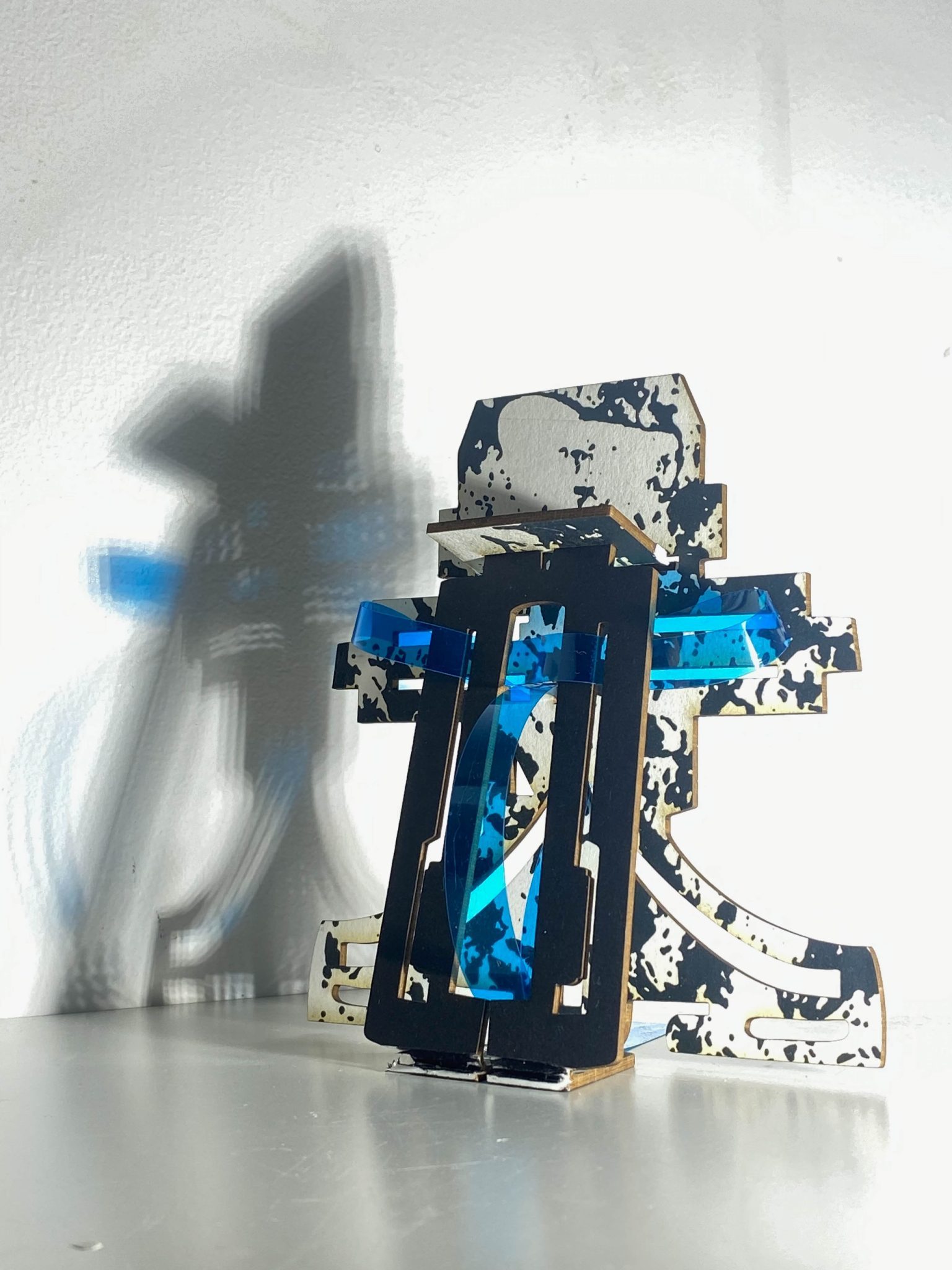 This small maquette is made from laser-cut, screen-printed card, cut and scored before being layered upright into a shallow architectural form, its front surrounded by curved wire mesh. Rectangular openings and a strip of blue film allow light to pass through, casting repeating, shifting shadows behind the work.