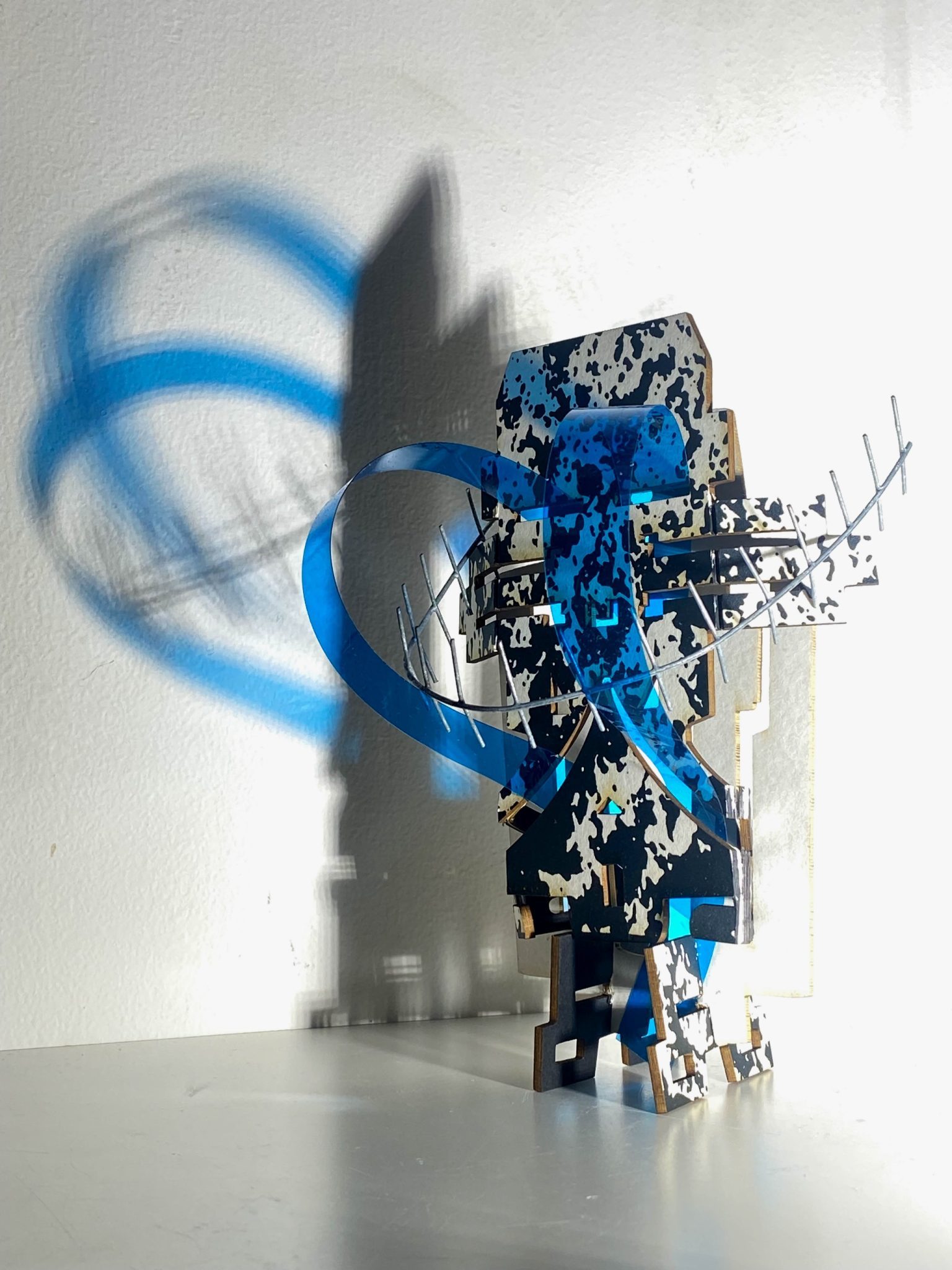 This small maquette is made from laser-cut, screen-printed card, cut and scored before being layered upright into a shallow architectural form, its front surrounded by curved wire mesh. Rectangular openings and a strip of blue film allow light to pass through, casting repeating, shifting shadows behind the work.