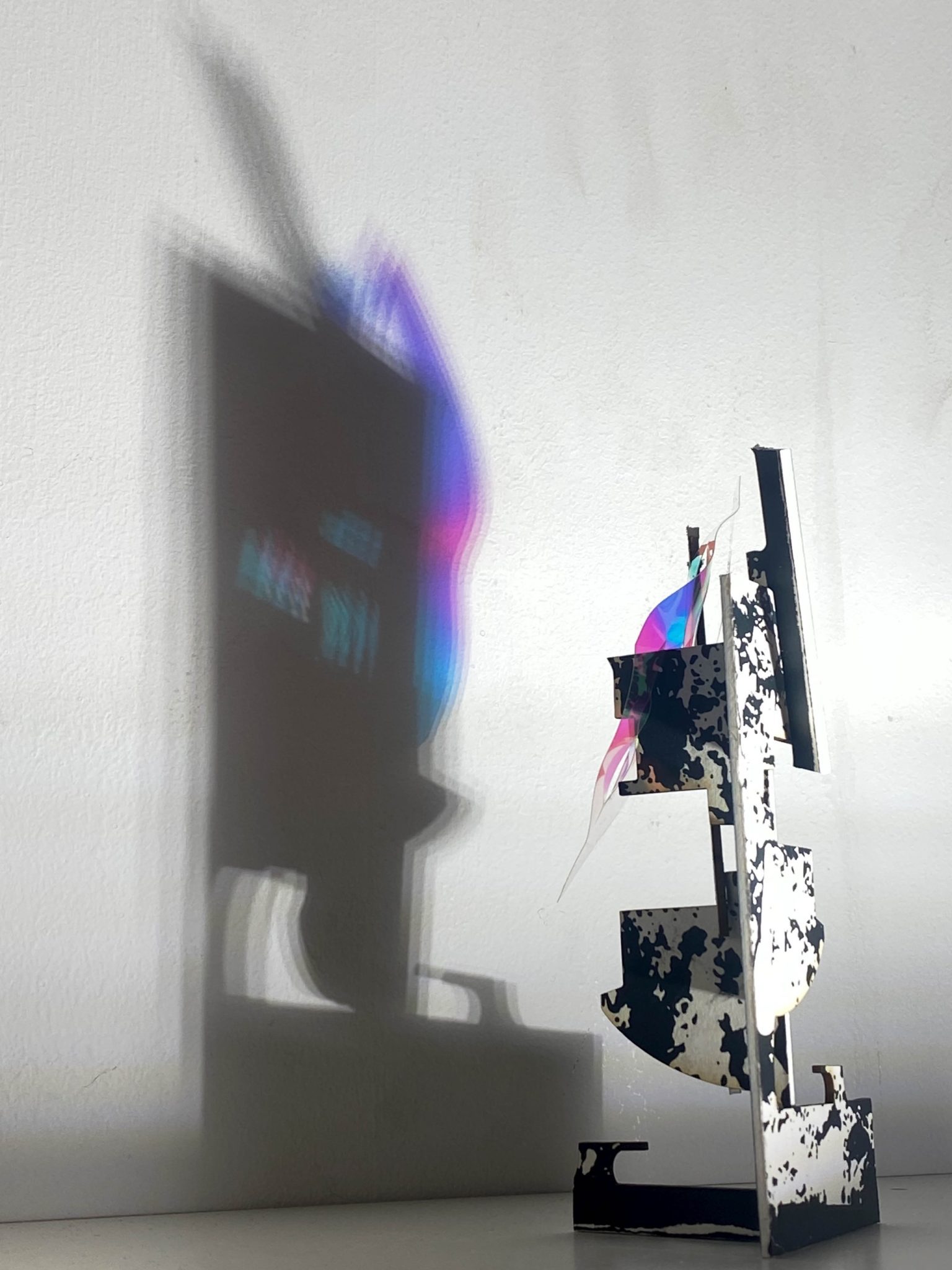 This small maquette is made from laser-cut, screen-printed card, cut and scored before being layered upright into a shallow architectural form. Rectangular openings and a strip of dichroic film allow light to pass through, casting repeating, colourful, shifting shadows behind the work.