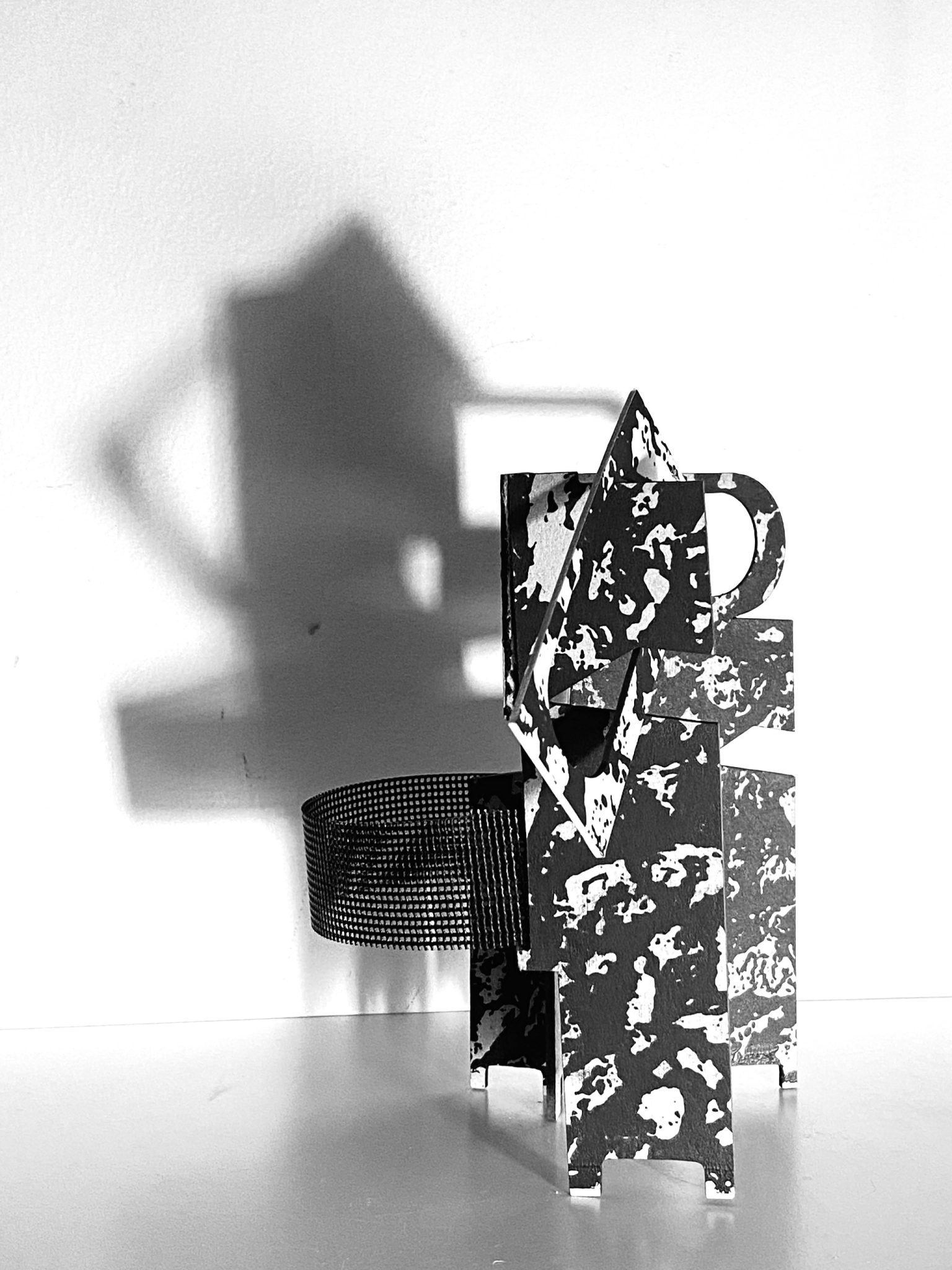 This small maquette is made from laser-cut, screen-printed card, cut and scored before being layered upright into a shallow architectural form. Rectangular openings and a strip of black mesh allow light to pass through, casting repeating, shifting shadows behind the work.