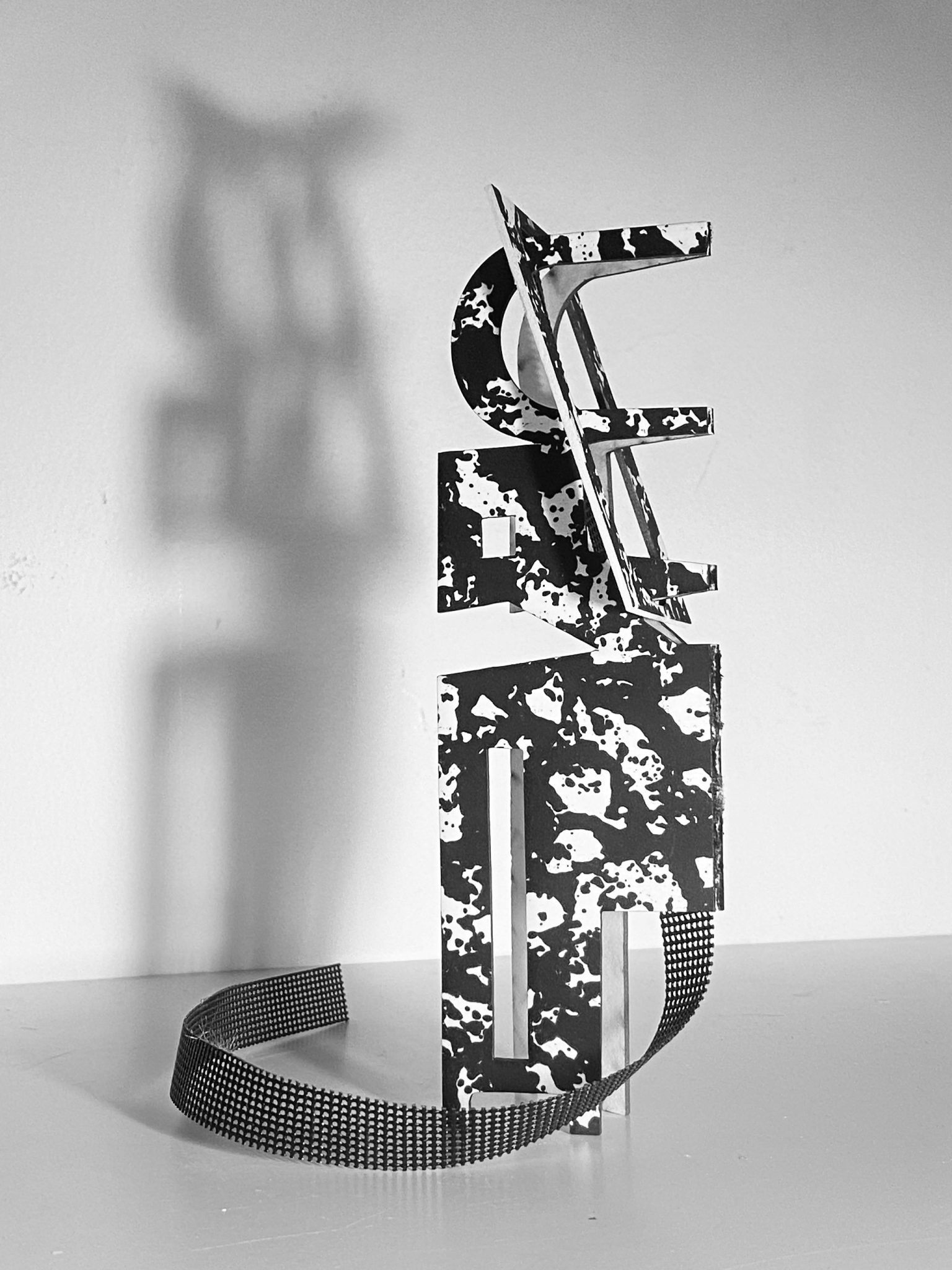 This work is a small maquette made from laser-cut card, presented upright on a flat surface. It is constructed from a flat screen printed card plane that is cut and scored, then layered vertically to form a shallow, architectural structure with rectangular openings. A strip of black mesh has been added. Light passes through the openings, creating overlapping shadows that repeat and shift the internal shapes onto the surface behind the work.
