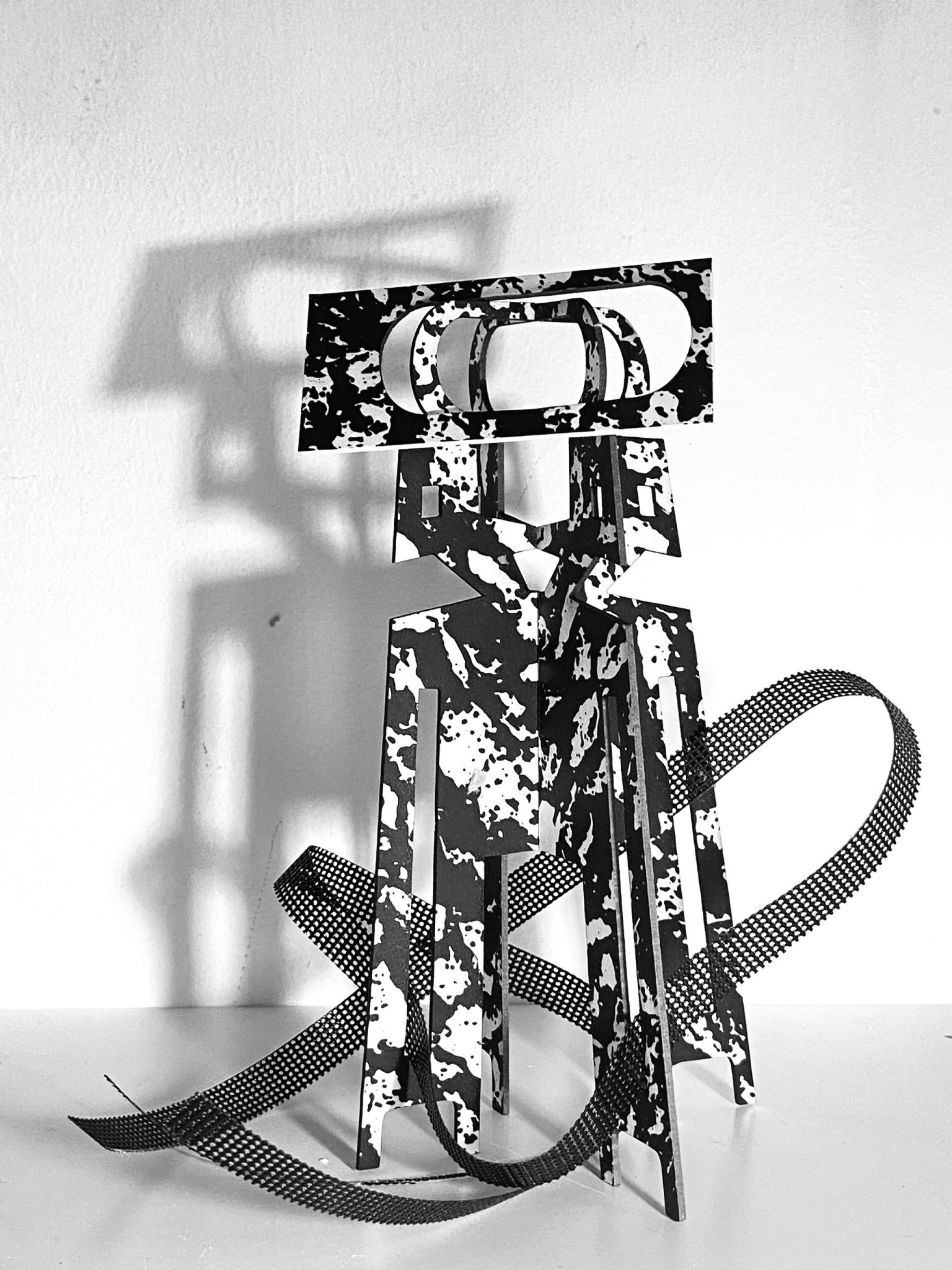 30 Day Maquette drill This work is a small maquette made from laser-cut card, presented upright on a flat surface. It is constructed from a flat screen printed card plane that is cut and scored, then layered vertically to form a shallow, architectural structure with rectangular openings. A strip of black mesh has been added. Light passes through the openings, creating overlapping shadows that repeat and shift the internal shapes onto the surface behind the work.