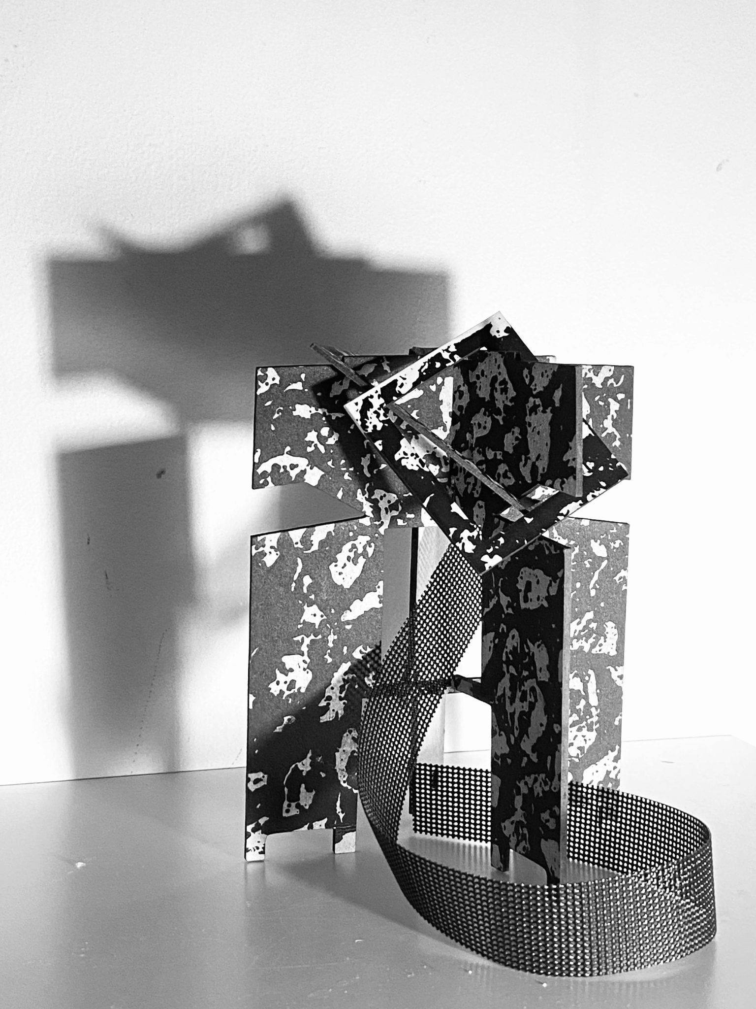 This small maquette is made from laser-cut, screen-printed card, cut and scored before being layered upright into a shallow architectural form. Rectangular openings and a strip of black mesh allow light to pass through, casting repeating, shifting shadows behind the work.
