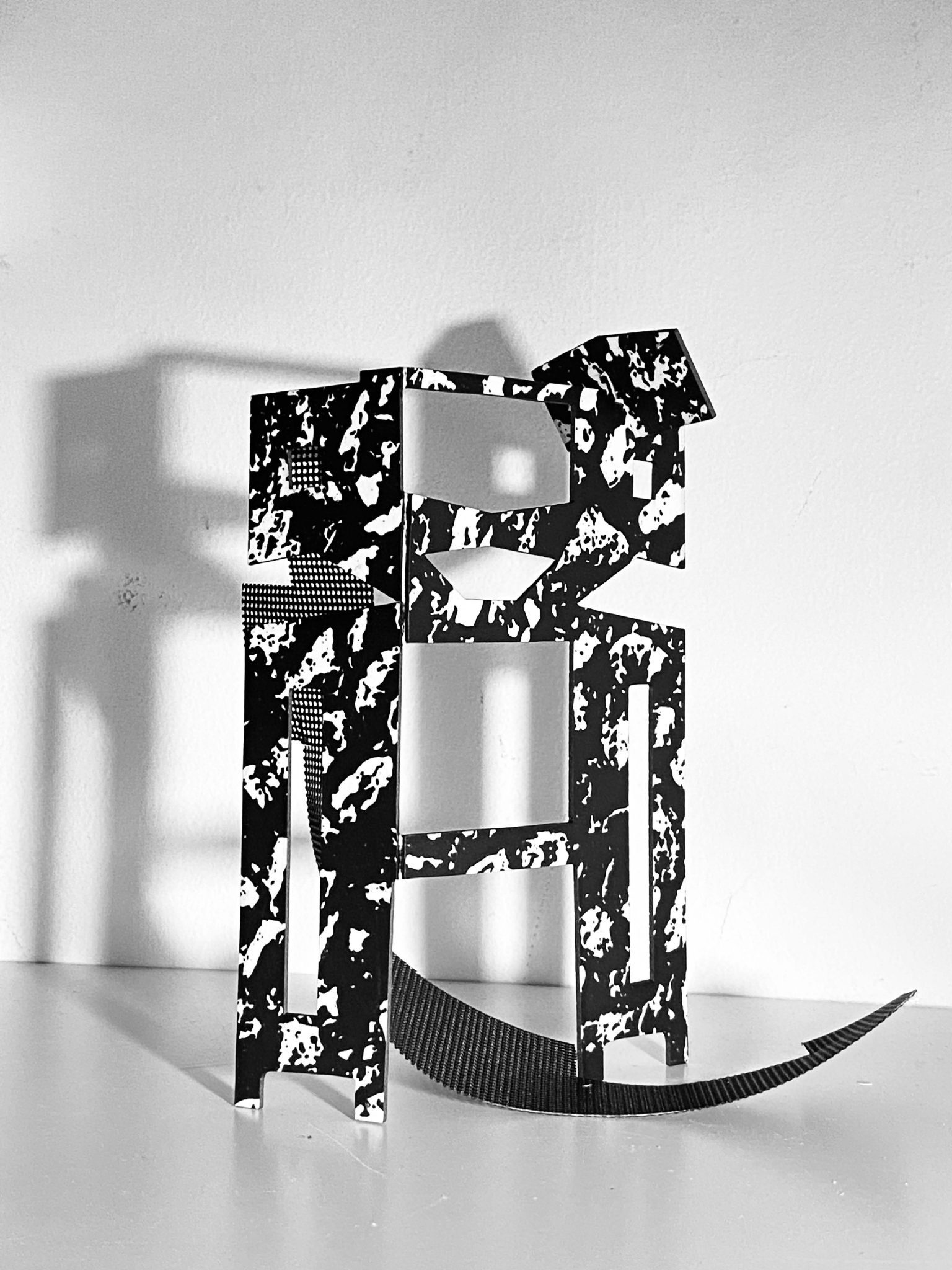 This work is a small maquette made from laser-cut card, presented upright on a flat surface. It is constructed from a flat screen printed card plane that is cut and scored, then layered vertically to form a shallow, architectural structure with rectangular openings. A strip of black mesh has been added. Light passes through the openings, creating overlapping shadows that repeat and shift the internal shapes onto the surface behind the work.