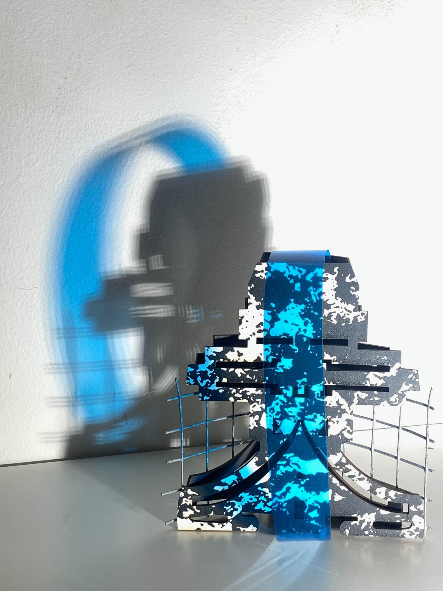This small maquette is made from laser-cut, screen-printed card, cut and scored before being layered upright into a shallow architectural form, its back surrounded by wire mesh. Rectangular openings and a strip of blue film allow light to pass through, casting repeating, shifting shadows behind the work.