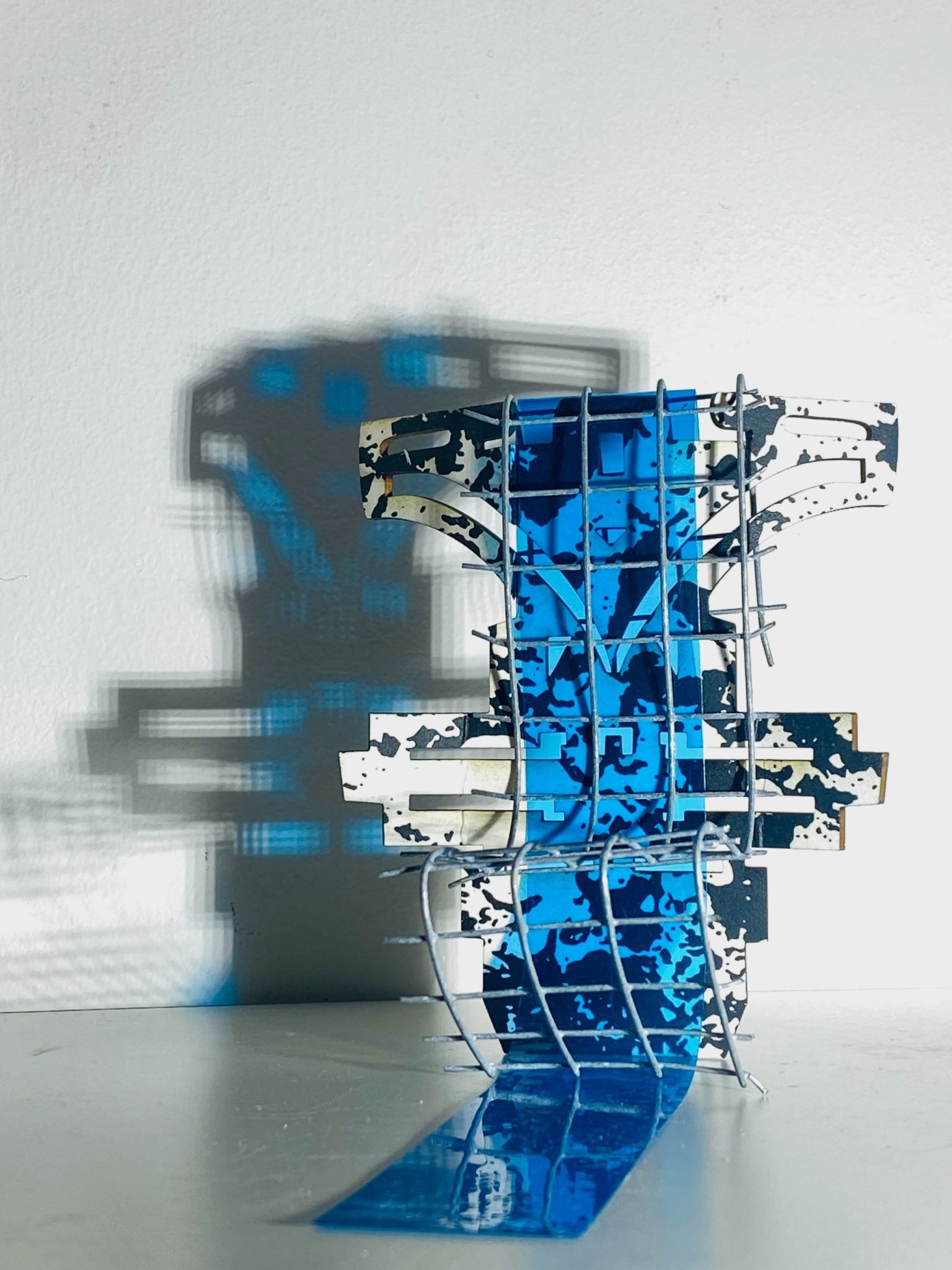 This small maquette is made from laser-cut, screen-printed card, cut and scored before being layered upright into a shallow architectural form, its front surrounded by curved wire mesh. Rectangular openings and a strip of blue film allow light to pass through, casting repeating, shifting shadows behind the work.