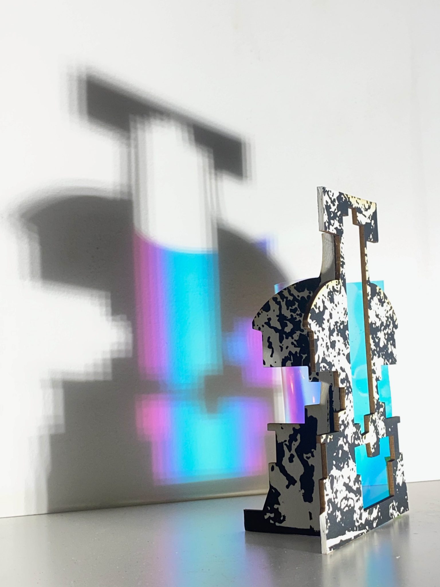 This small maquette is made from laser-cut, screen-printed card, cut and scored before being layered upright into a shallow architectural form. Rectangular openings and a strip of picric film allow light to pass through, casting repeating, colourful, shifting shadows behind the work.