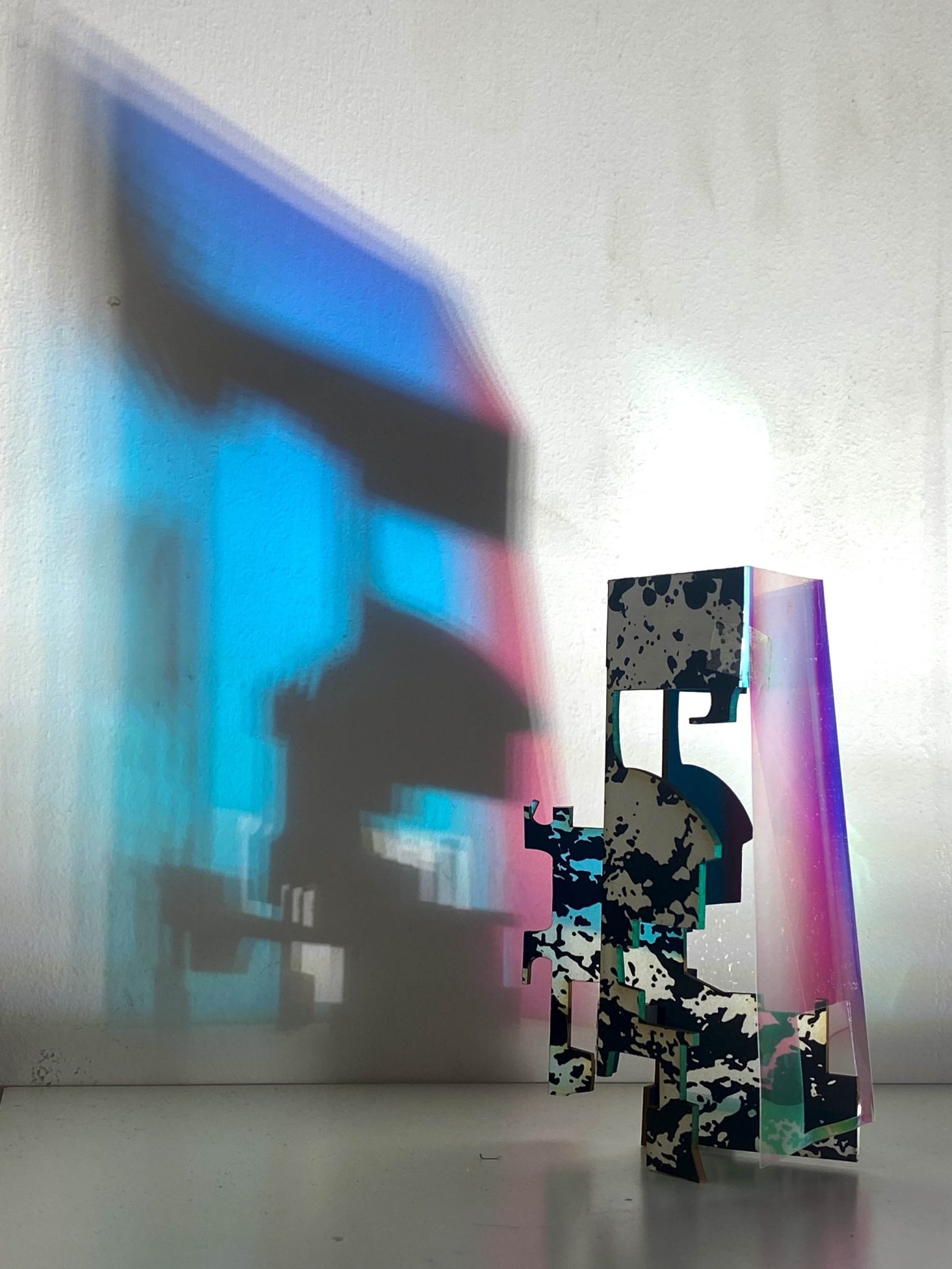 This small maquette is made from laser-cut, screen-printed card, cut and scored before being layered upright into a shallow architectural form. Rectangular openings and a strip of dichroic film allow light to pass through, casting repeating, colourful, shifting shadows behind the work.