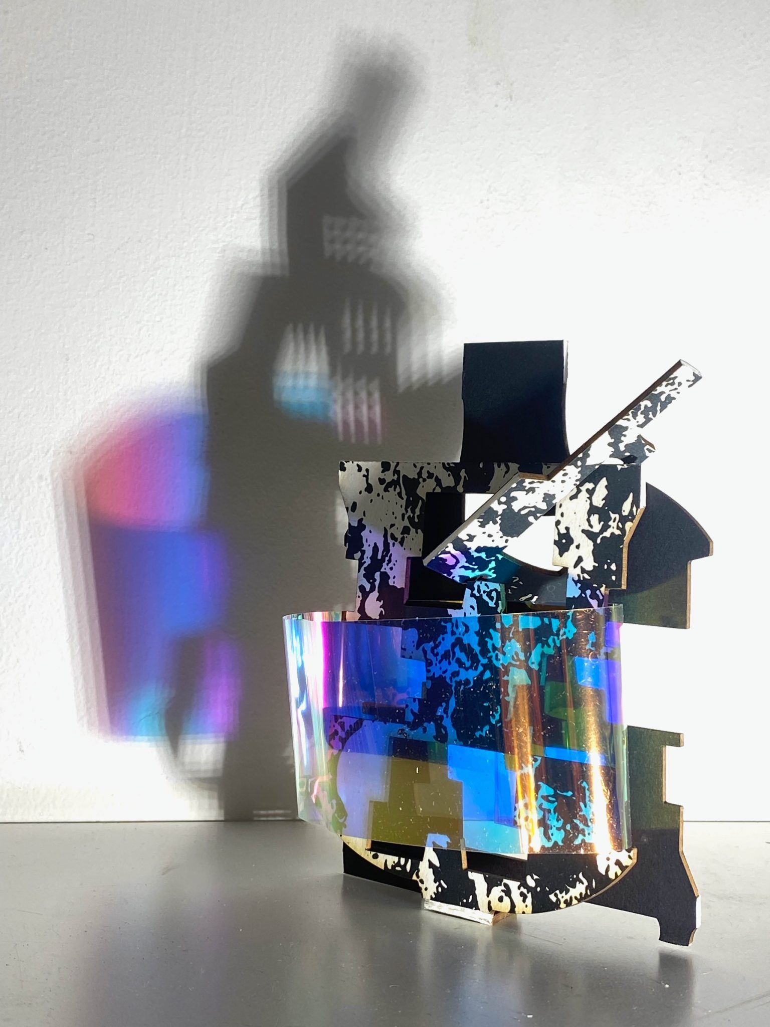 This small maquette is made from laser-cut, screen-printed card, cut and scored before being layered upright into a shallow architectural form. Rectangular openings and strips of picric film allow light to pass through, casting repeating, colourful, shifting shadows behind the work.