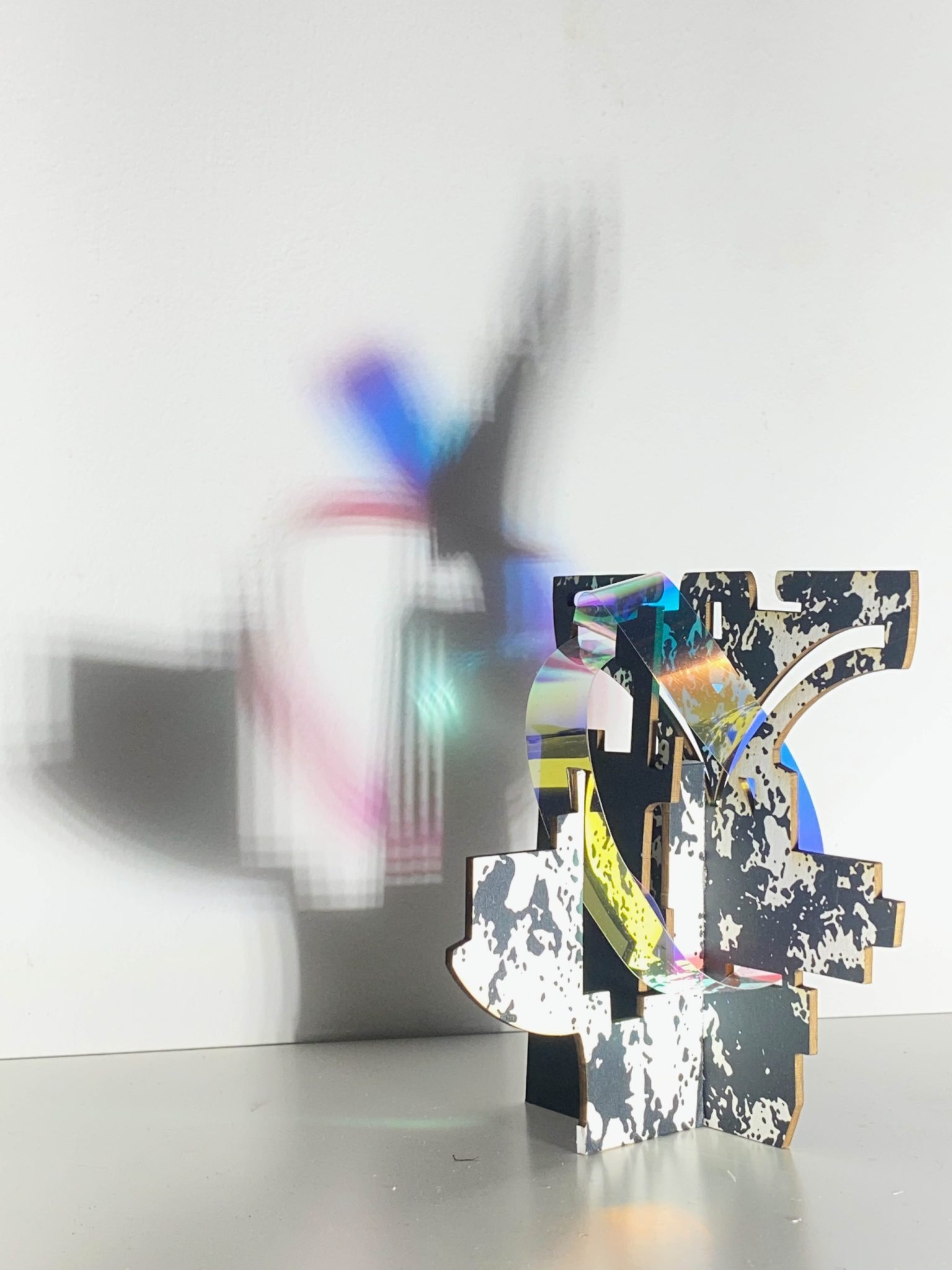 This small maquette is made from laser-cut, screen-printed card, cut and scored before being layered upright into a shallow architectural form. Rectangular openings and a strip of dichroic film allow light to pass through, casting repeating, colourful, shifting shadows behind the work.