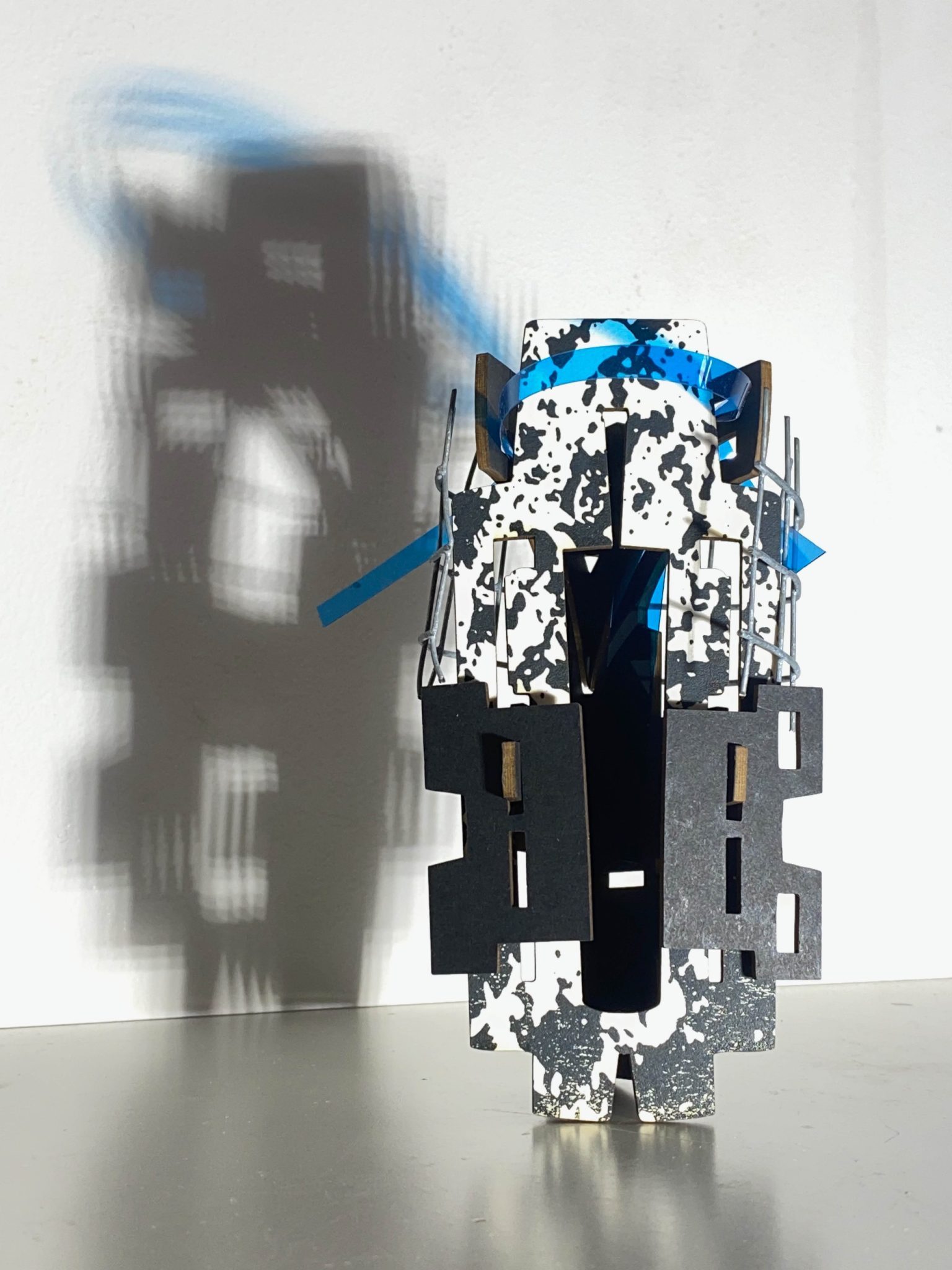 This small maquette is made from laser-cut, screen-printed card, cut and scored before being layered upright into a shallow architectural form, its front surrounded by curved wire mesh. Rectangular openings and a strip of blue film allow light to pass through, casting repeating, shifting shadows behind the work.