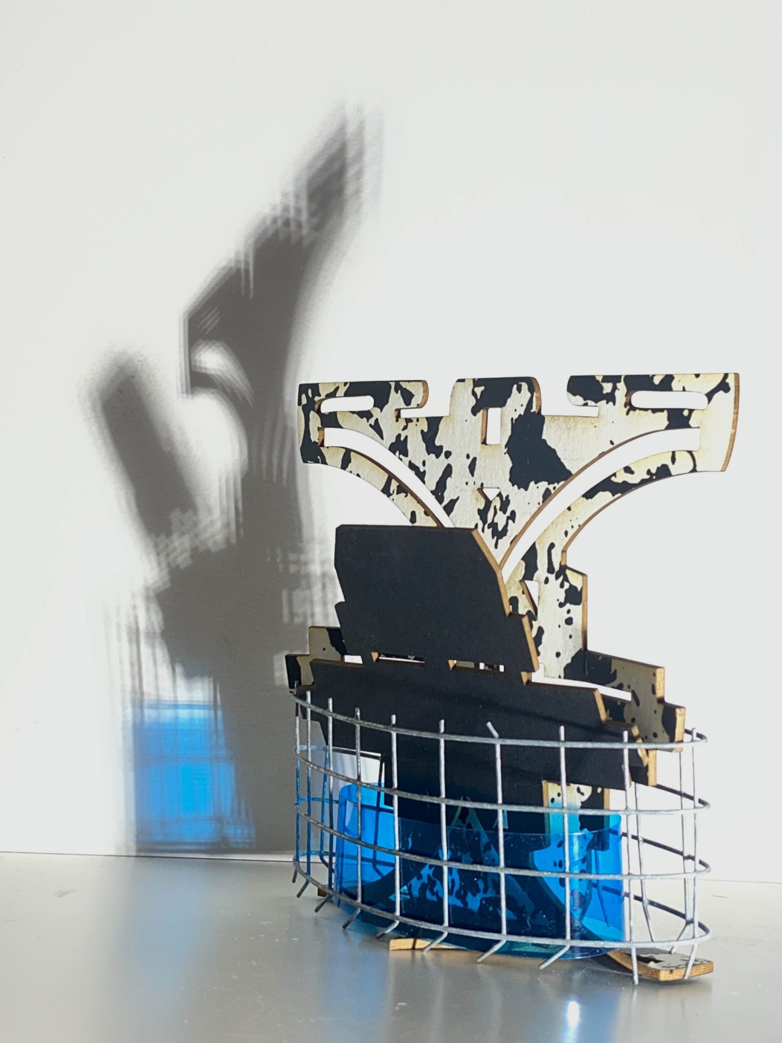 This small maquette is made from laser-cut, screen-printed card, cut and scored before being layered upright into a shallow architectural form, its front surrounded by curved wire mesh. Rectangular openings and a strip of blue film allow light to pass through, casting repeating, shifting shadows behind the work.