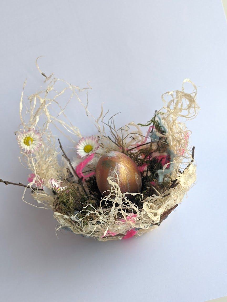 The nest is made from twigs, moss, and fine natural fibres holding a single metallic green‑and‑gold painted egg. Small pink and white flowers are arranged around the egg.