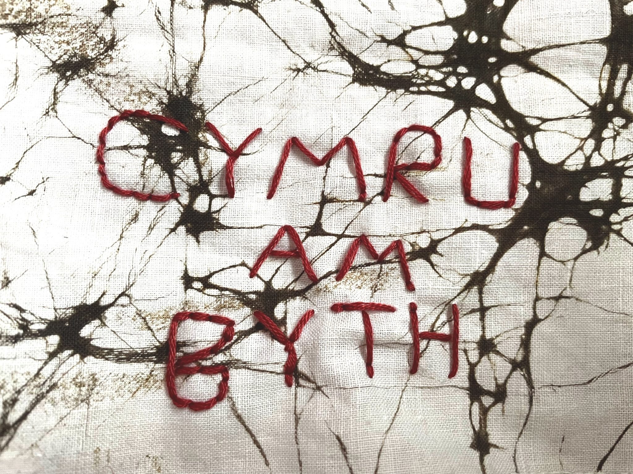 fabric background, white fabric with black ink blots. stitched on top in red: Cymru am byth.