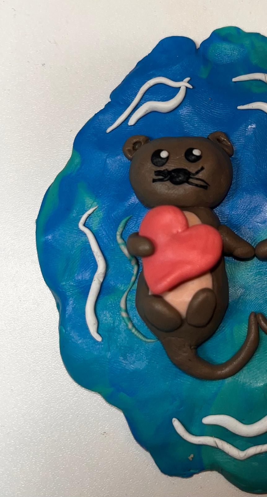 An otter made out of clay - holding a big heart. Floating on water