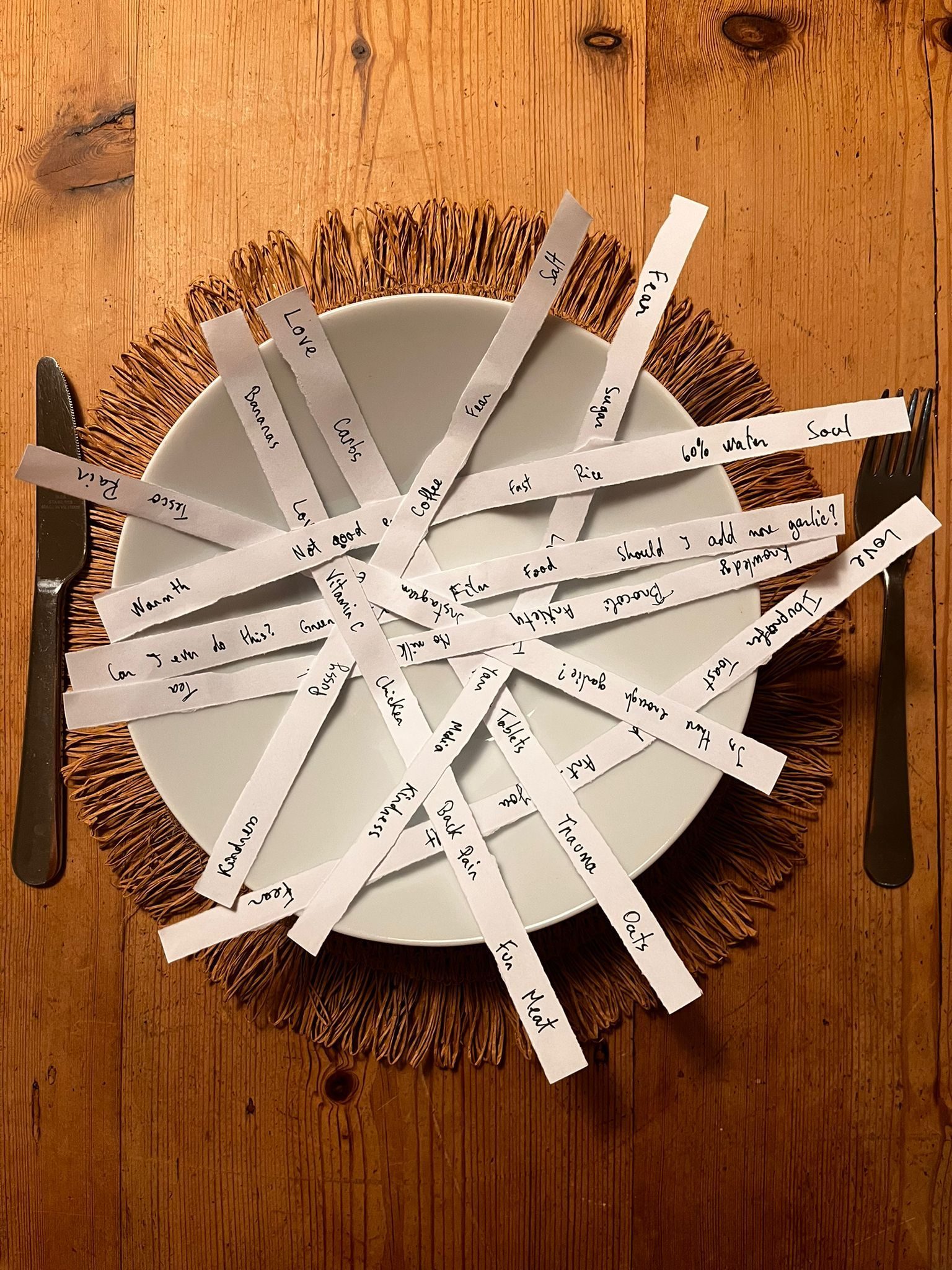 Strips of paper with words written on them 'Fear, Love, Ibuprofen, Trauma, Sugar, Heart'  - all placed in a bowl on the table with a fork and a knife side by side.