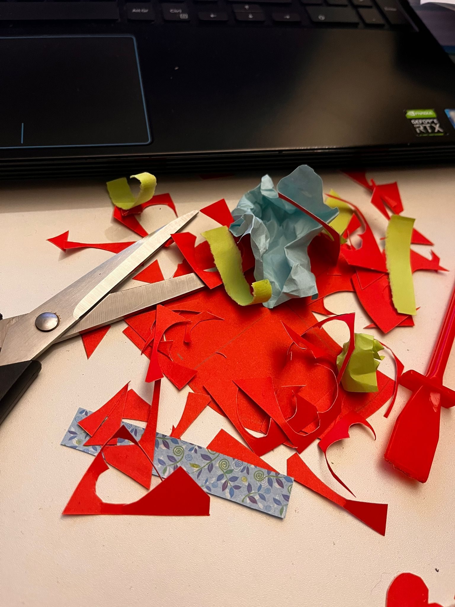 A pile of coloured paper scaraps left from crafting someting. On the side there's a pair of scissors, a glue stich and at hte back it the keypad of a laptop sugessting something else is going on at the same time as crafting