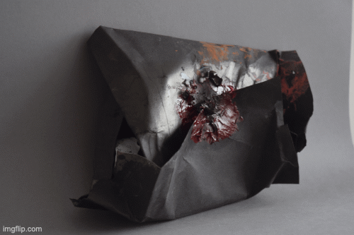 A black sculpture in a parallelogram shape sits in front of a grey backdrop. There are paint marks in orange and pink on the envelope in two places and what looks like a paint seal in crimson in the centre, covering the meeting of one side of the paper on top of the other.