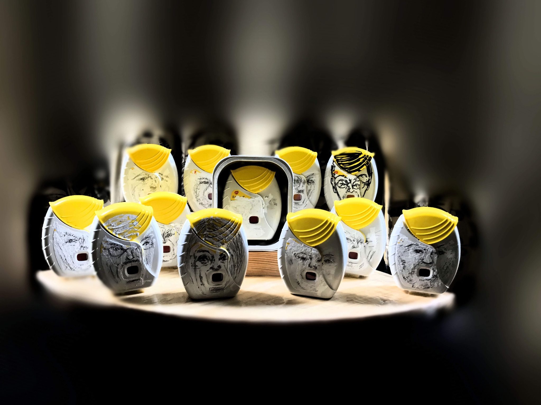 a collection of objects that are probably asthma inhalers or similar devices, arranged with one that has no given features, they have the information on labelling obscured by tipped and pencilled faces that could be such people as `John `north, Steve e Bannon, is this a coincidencei? `probably not? they are arranged on a wooden matrix and the lighting is dramatic over hyped and threatening