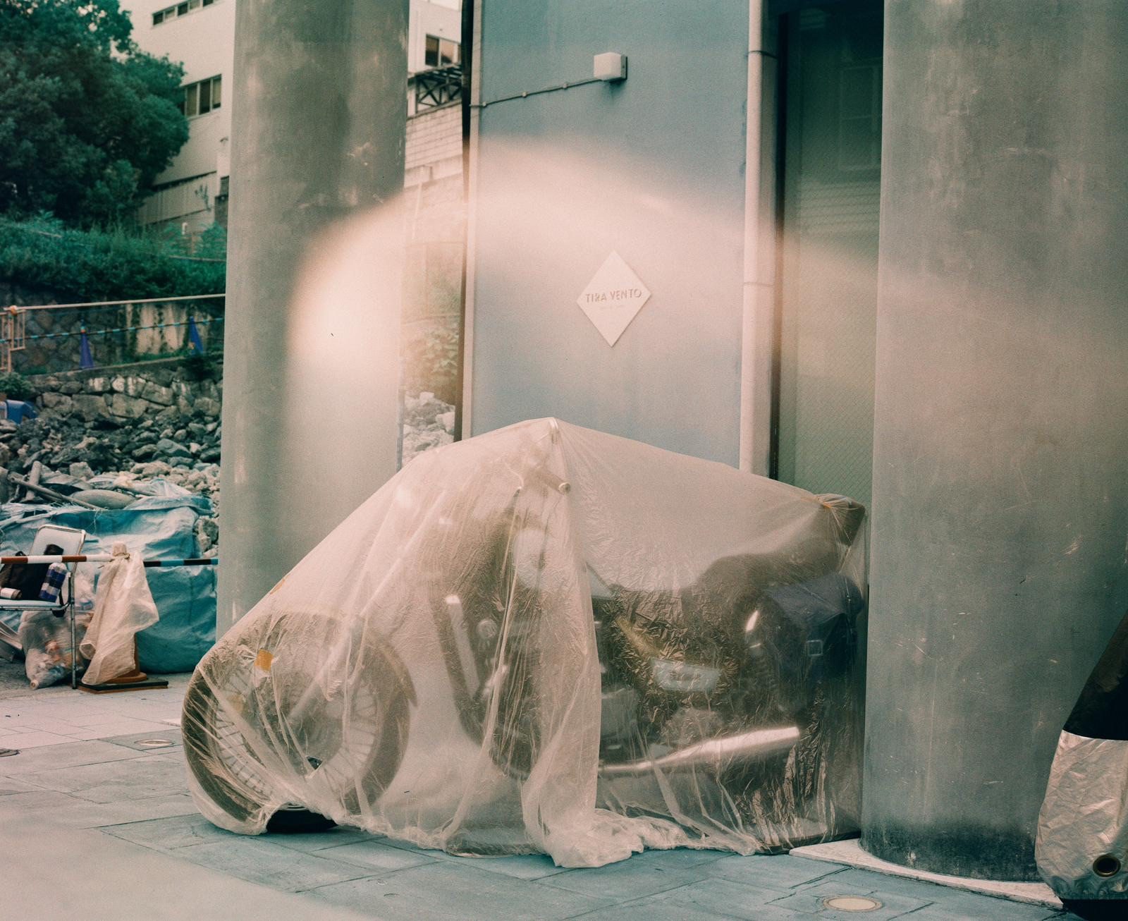 A photograph of a motorbike covered with a thin plastic sheet to protect it from dust. It is shot on turquoise film so has a strange blue filter. The camera had a light leak so there is a patch of light in the center of the image that happens to echo the shape of the motorcycle.