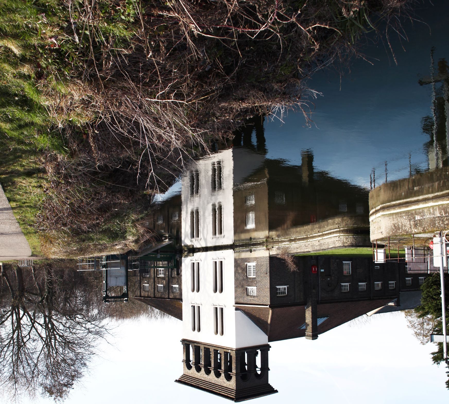 a photograph showing a brown brick building structure with a white tower, reflected in a water surface, but it's the reflection that is more prominent and at the top of the picture, and the real building serves as its reflection