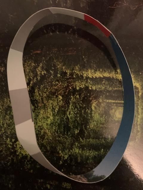 Colour photo of a Mobius strip, grey/white/red/blue irregular stripes curving into red rock formation layers as the strip turns. The strip sits on a shiny green surface which has a maze-like pattern on it. Shadowy indistinct head and shoulders are reflected in the shiny surface; so is a clear darker curve.