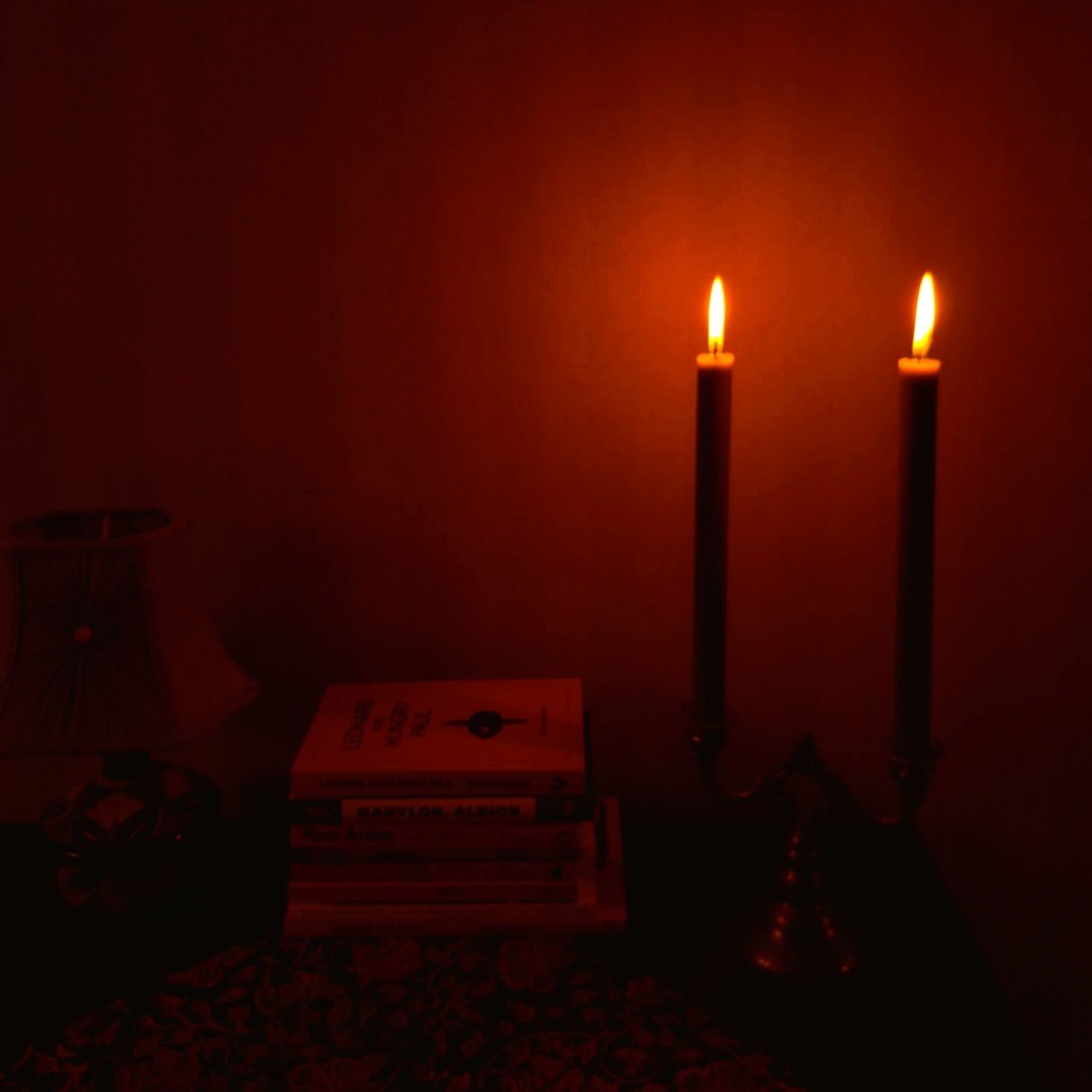 A photo of two candles burning in a dark room. The candles give off a reddish glow and there's a pile of books on the table.
