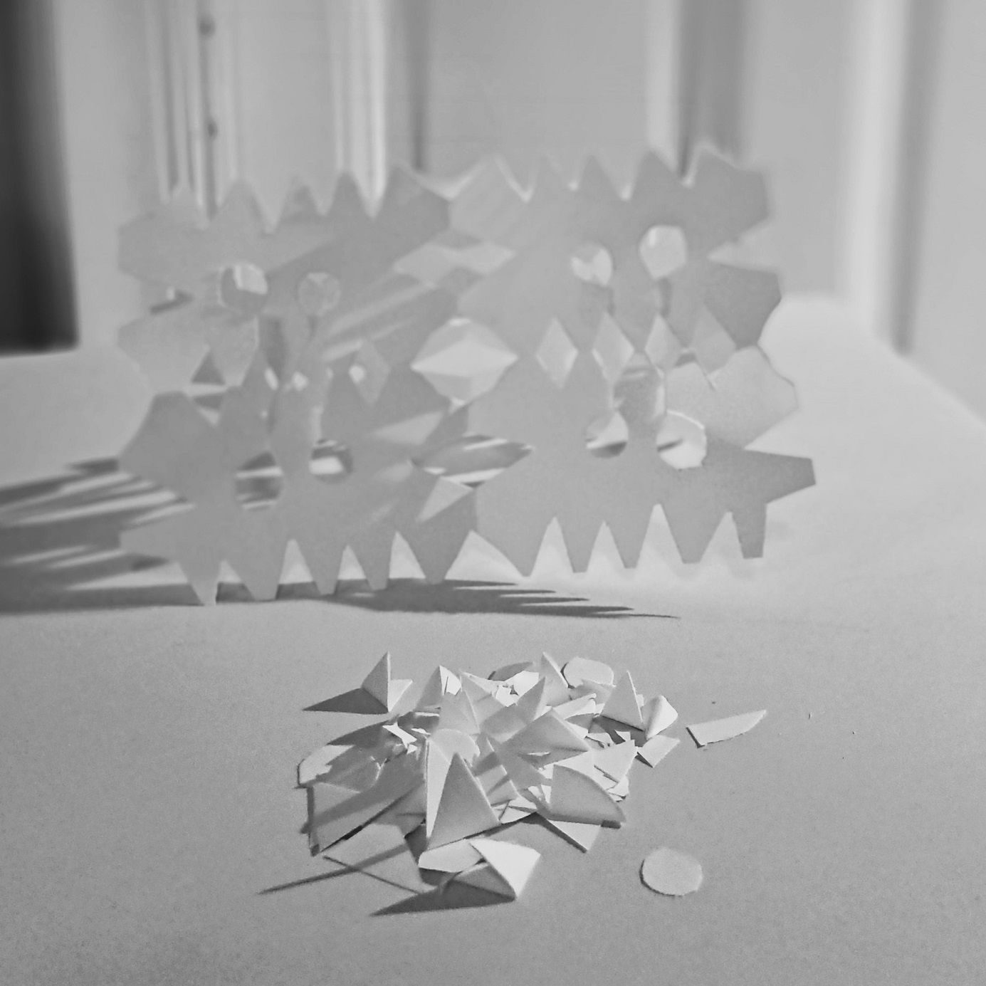 In the foreground, paper cuttings from making paper art. In the background, a piece of folded out paper with cut out shapes resembling a doily.