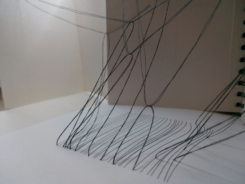 abstract drawing with black thread and black pen, view from the ground, threads emerging out of the drawing and connecting to pages of concertina sketchbook, some going to the left, some to the right, some looser than others