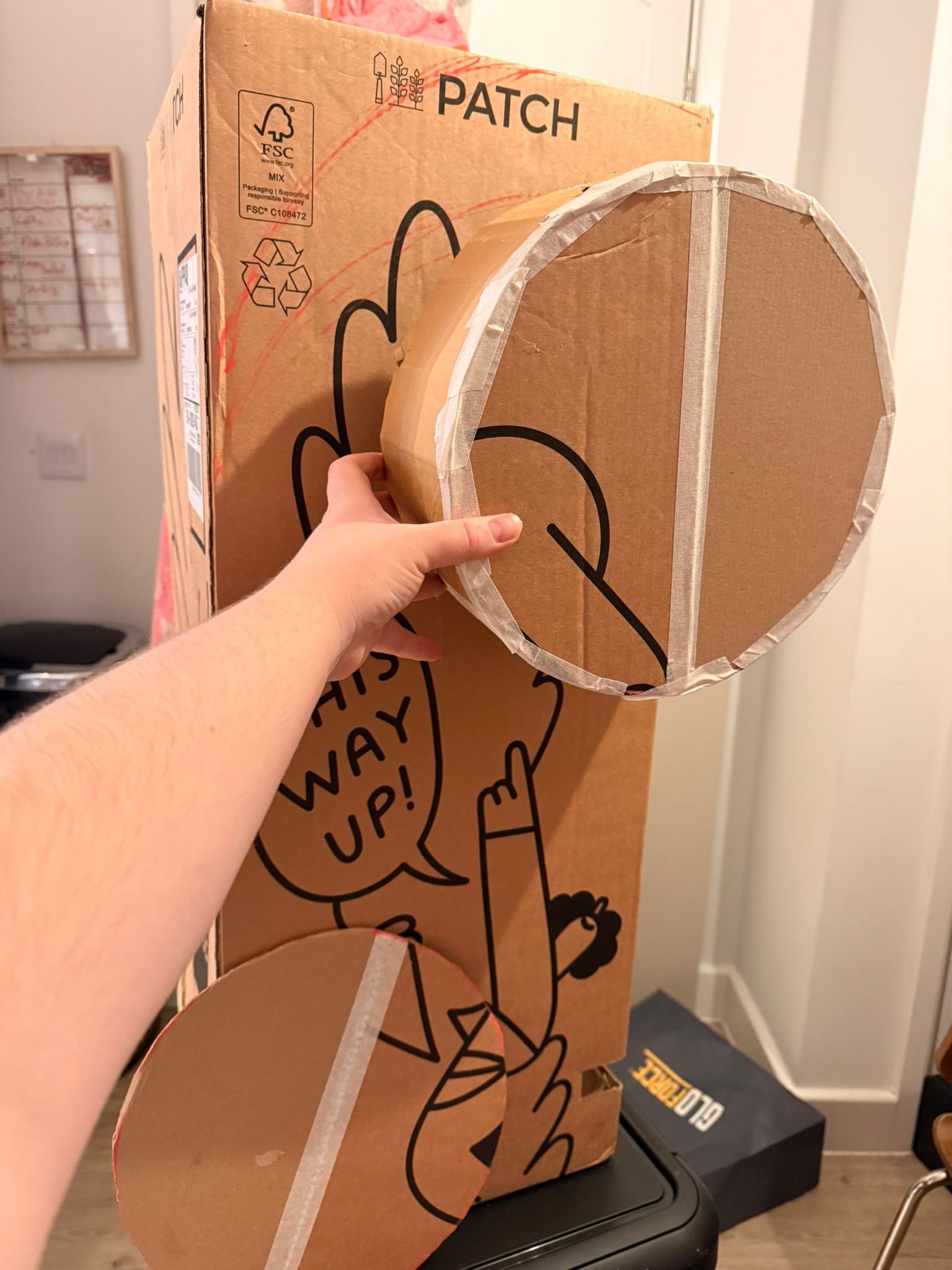 Photo of a tall cardboard box, with a hand holding up another pieced together 3d circle box to the side. There are some scribbles in red on the taller box too. as well as a little circle cut out resting against it.