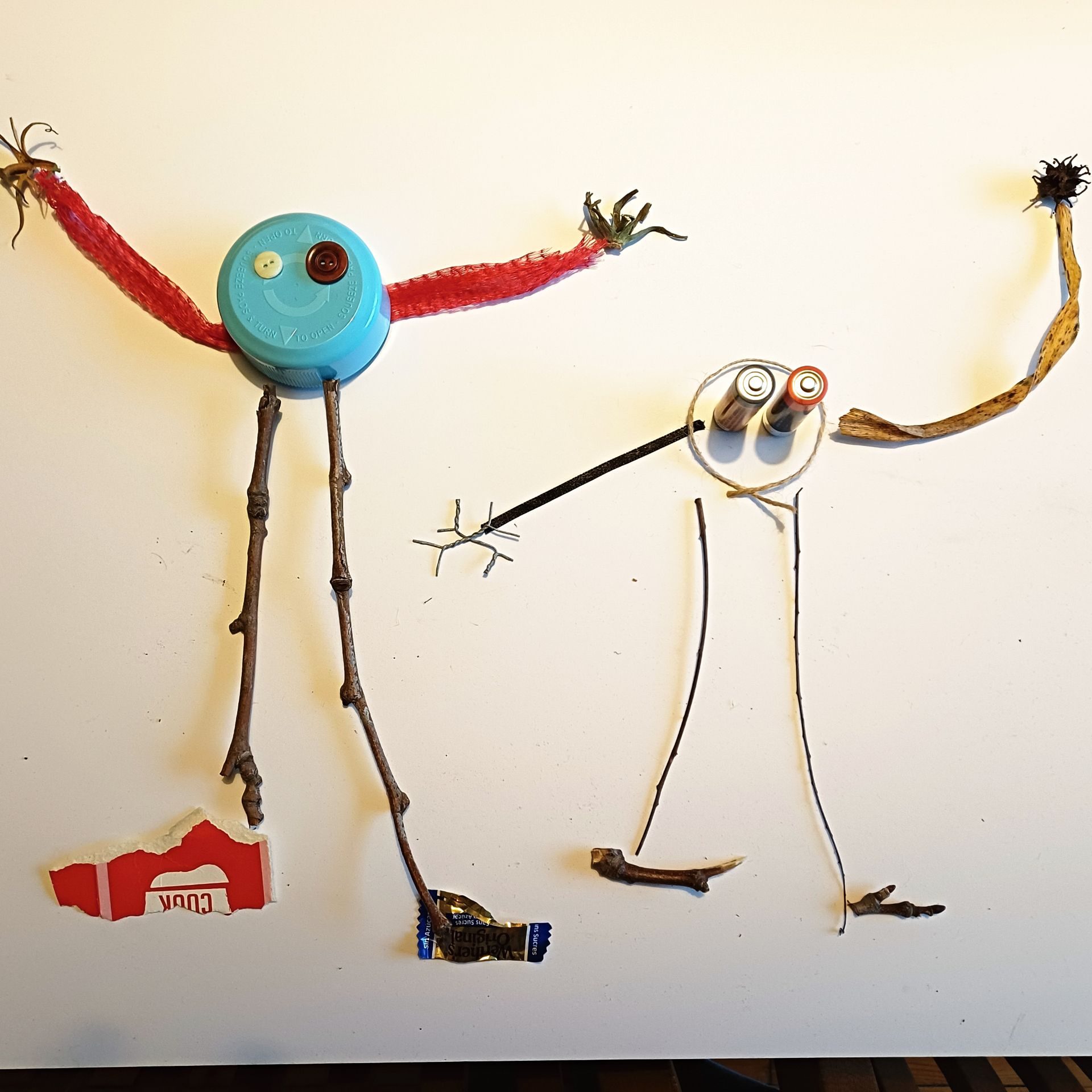 2 simplified figures are drawn by placing ad hoc objects like buttons, batteries, twigs etc to resemble children’s first drawings, ie arms and legs coming off the head.  The work appears absurd and humorous with a manic presence.