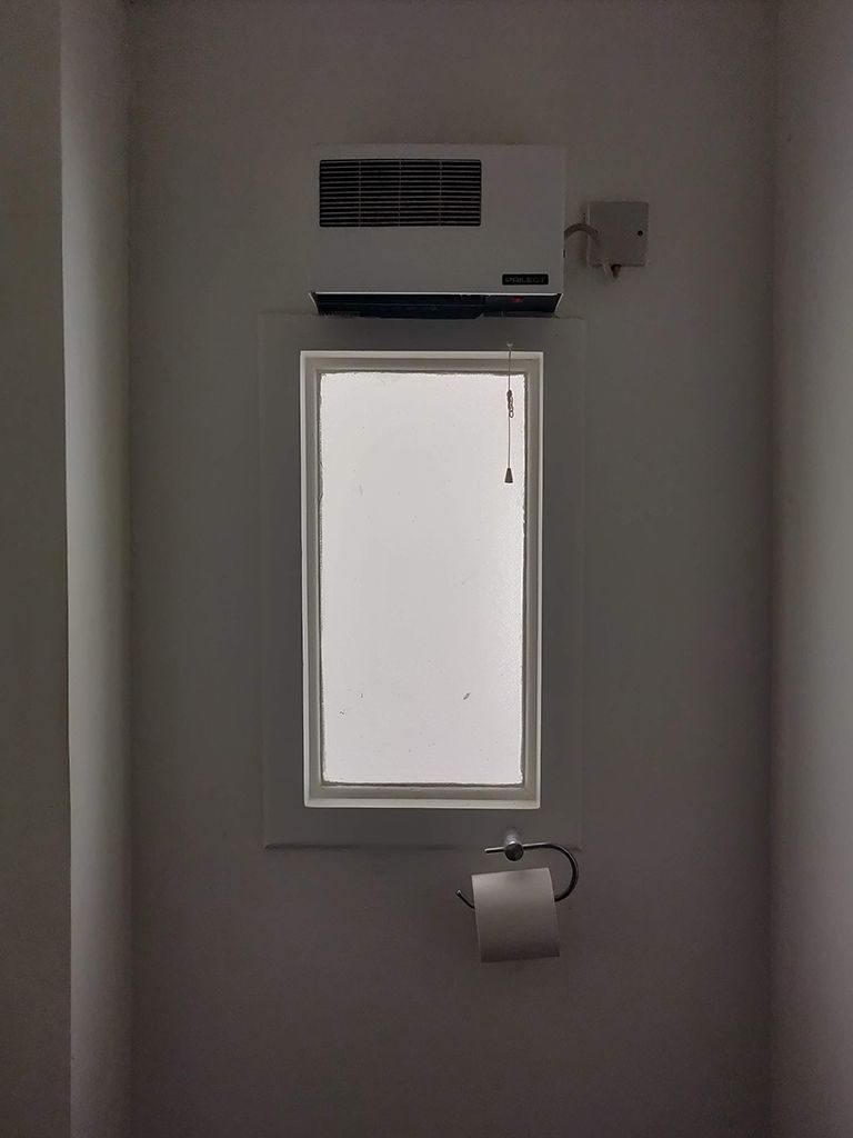 A colour photograph of a small bathroom window with a roll of toilet paper and extractor fan set in a plain white painted alcove