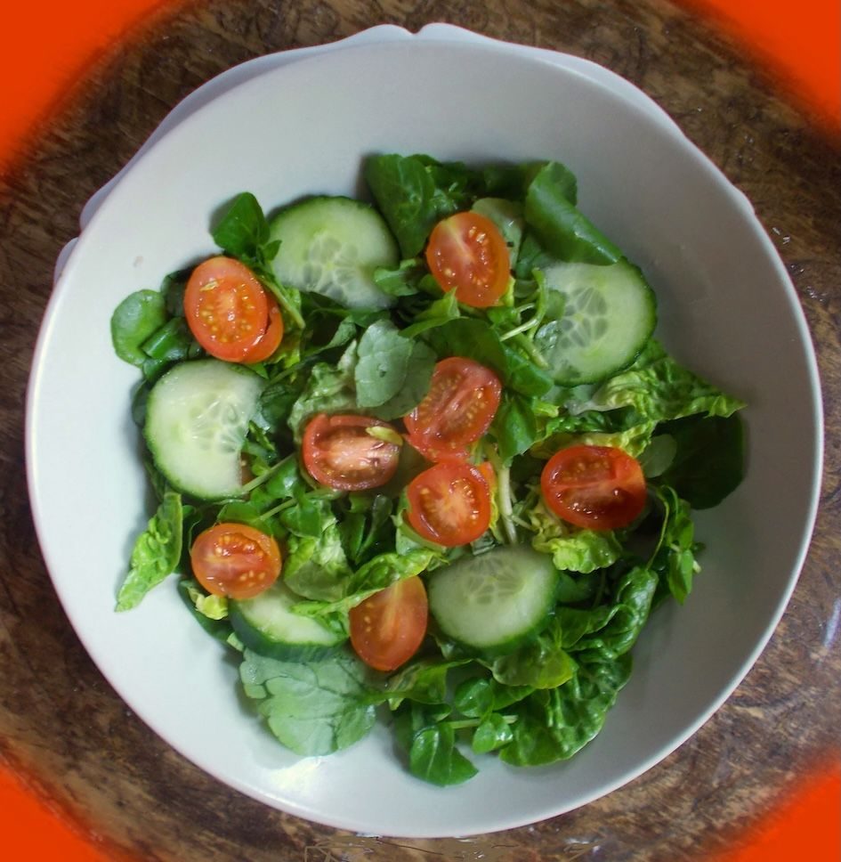 photograph of a salad comprised of greens and red tomatoes, placed in circle white bowl on brown and red background which is square
