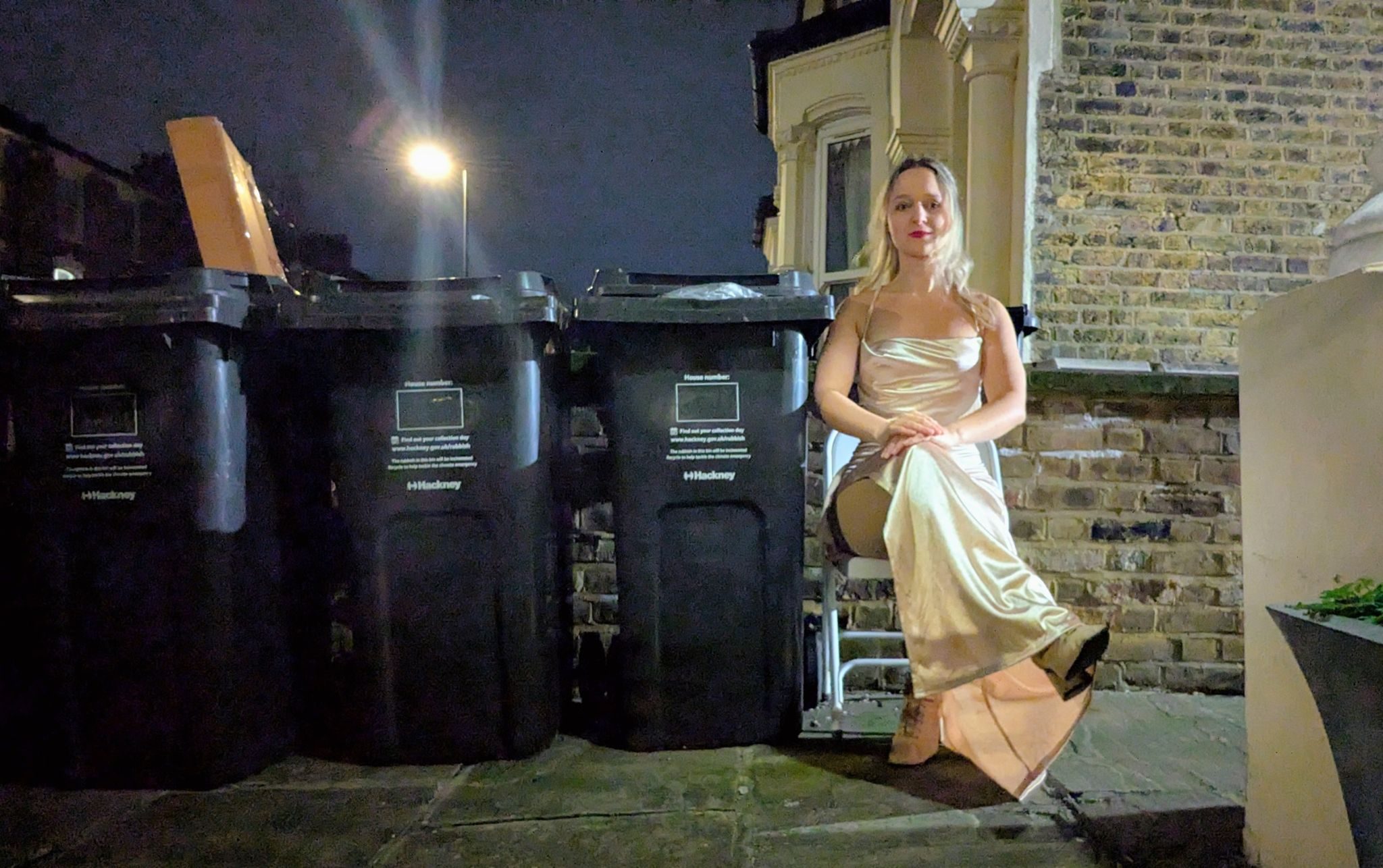 a blonde female presenting human in a long creme coloured ball gown sitting on a chair, next to 3 bins, in the dark, outside