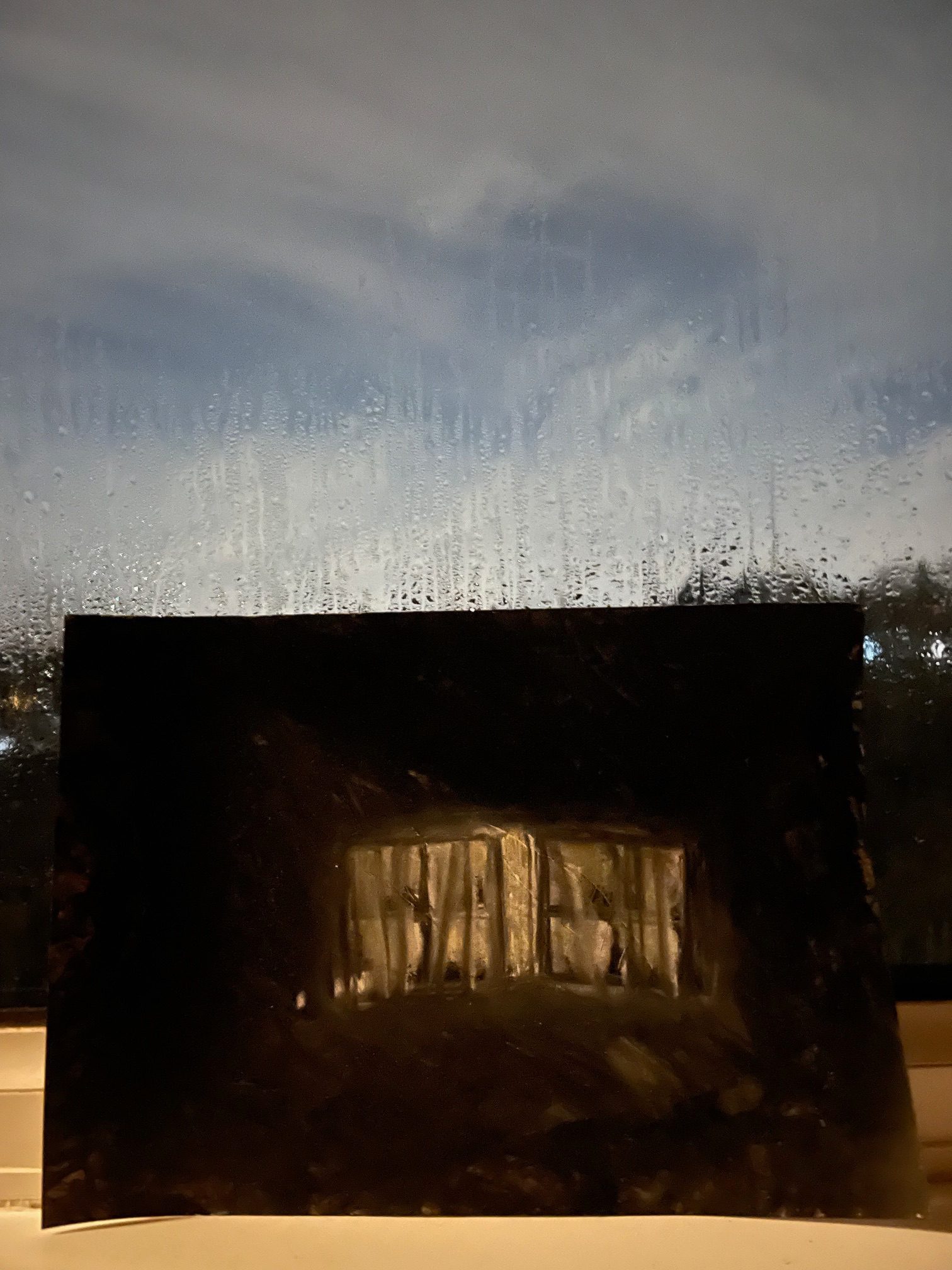 A charcoal and black acrylic paint drawing of a slightly opened window in the darkness. The A4 picture is landscape and photographed on the windowsill of a window. It is dark outside and rain drops are spattered on the glass