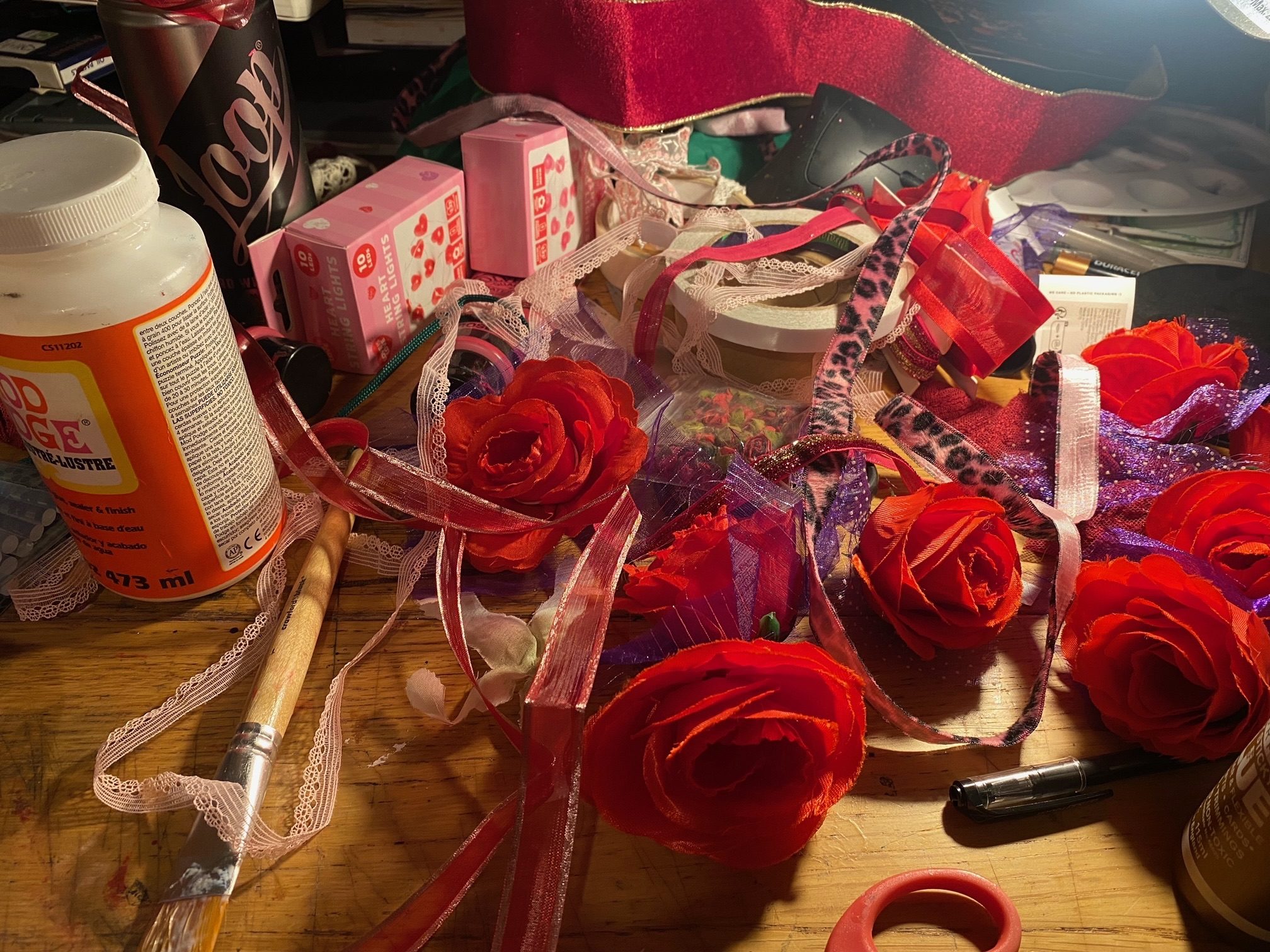 A desk covered I. Ribbon, fake flowers, paint brushes, glue and other craft bits