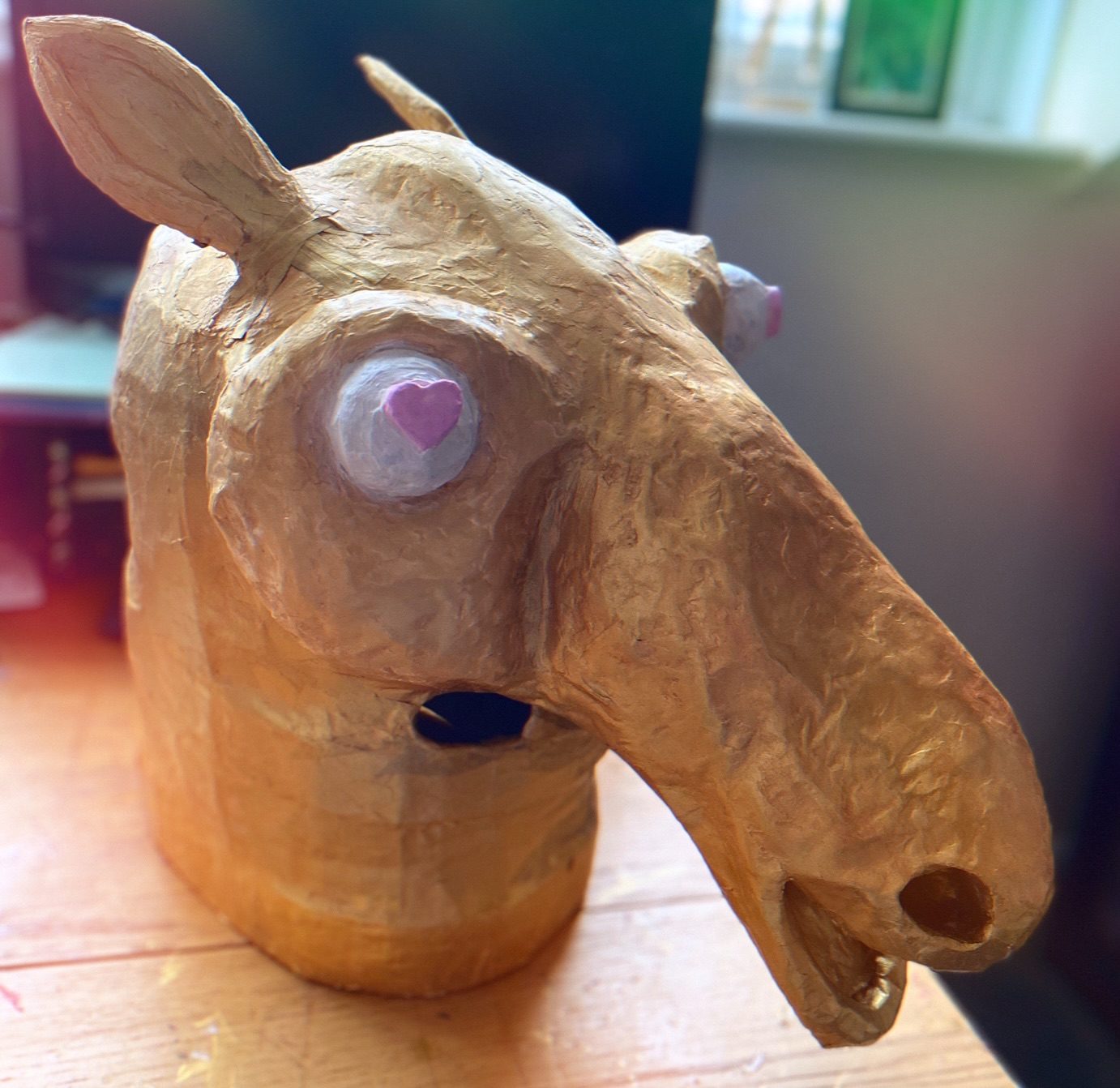 A paper mache mask of a carousel horse’s head, painted gold with a white eyeball with a pink loveheart in the centre
