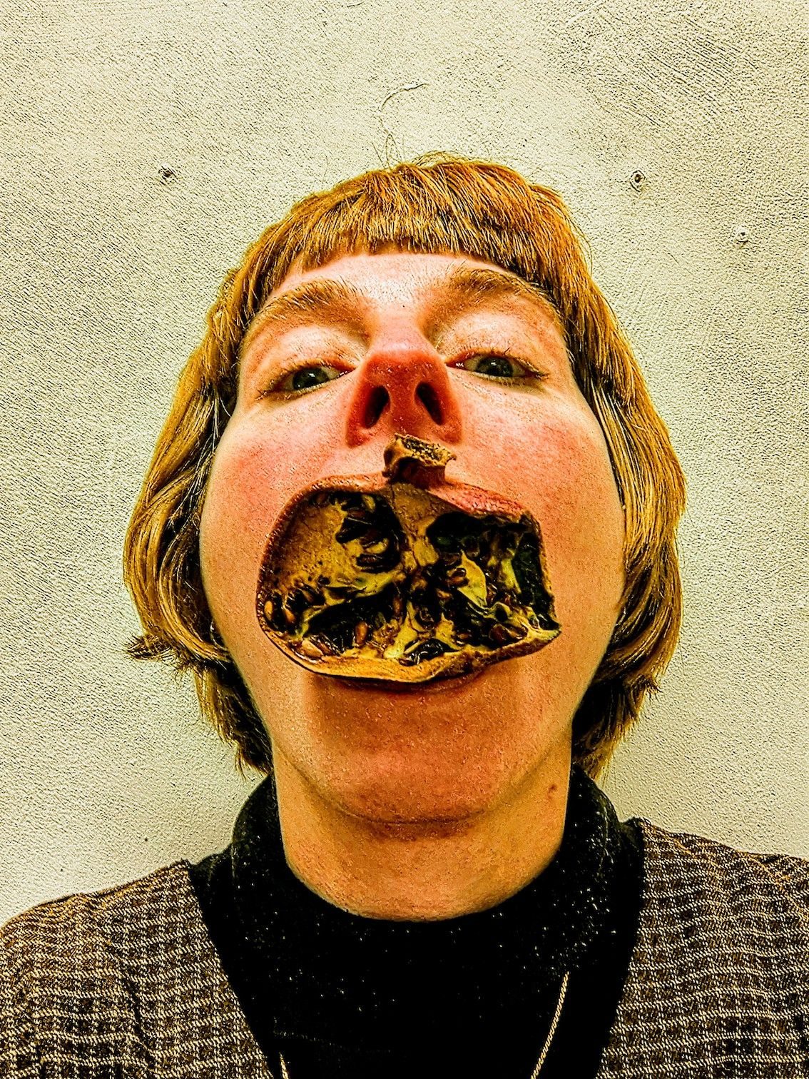 Image of the artists face, tilted upright with a dry half of a pomegranate in their mouth at a funny angle, the framing is like a passport photo.
