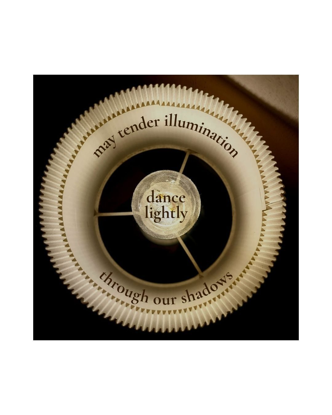 a close up photo of a light shot from above with a poem interwoven that reads: may tender illumination dance lightly through our shadows