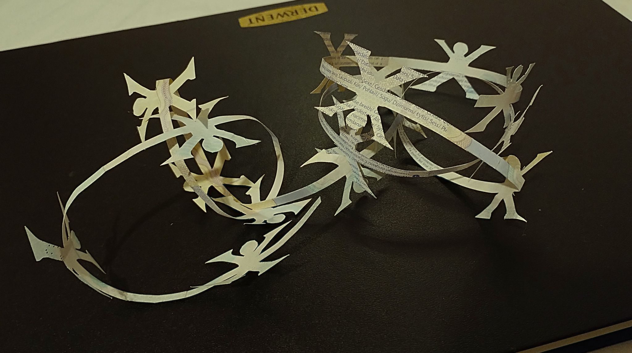 The Cost of Living is a tiny sculpture made from the inner paper pages of a UK passport. It comprises multiple figures arranged in a paper chain , linked together in a long strand made to look like barbed wire.