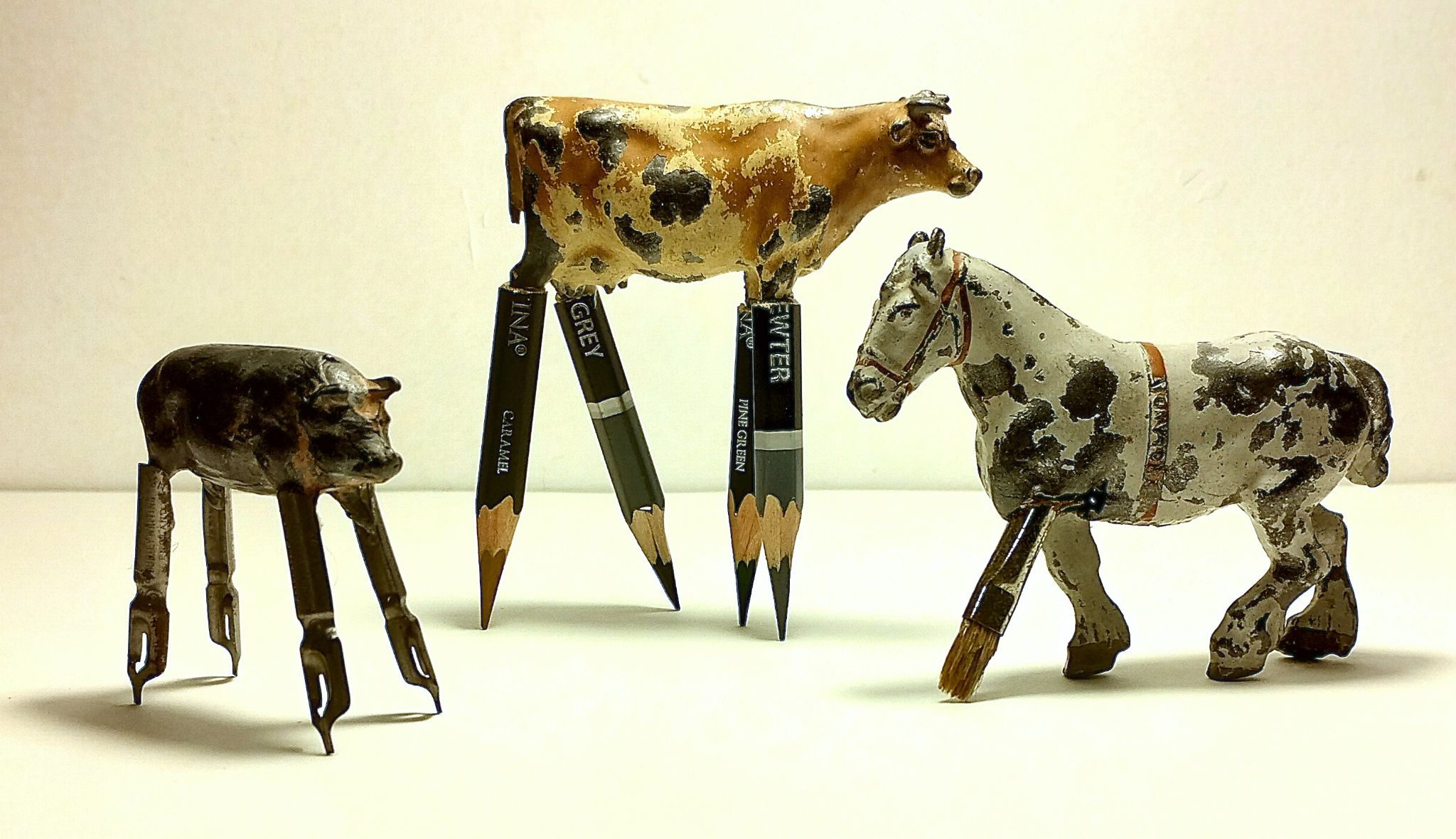 A collection of three small sculptures made from lead farm animals, given legs of pencils, pen nibs and a paintbrush