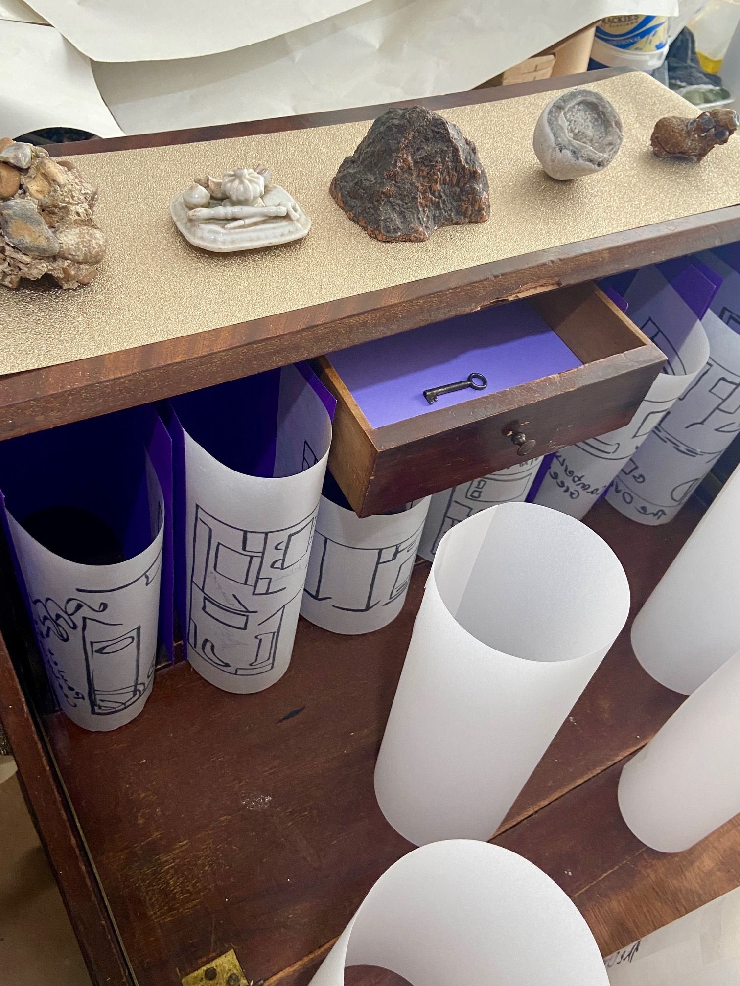 a bureau containing cylindrical floor plans; a key in small open drawer; a series of objects on the top of the bureau